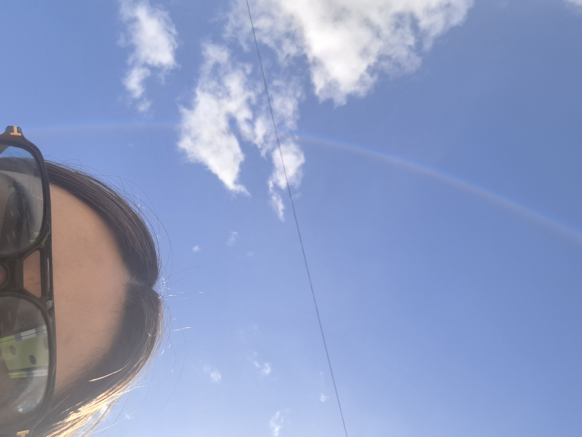 The image shows a partial view of a person with dark hair and sunglasses. The background is a bright blue sky with white clouds and a rainbow. There is also a thin wire running diagonally across the sky. There is no information provided in the image about Chaikovsky, Russia.