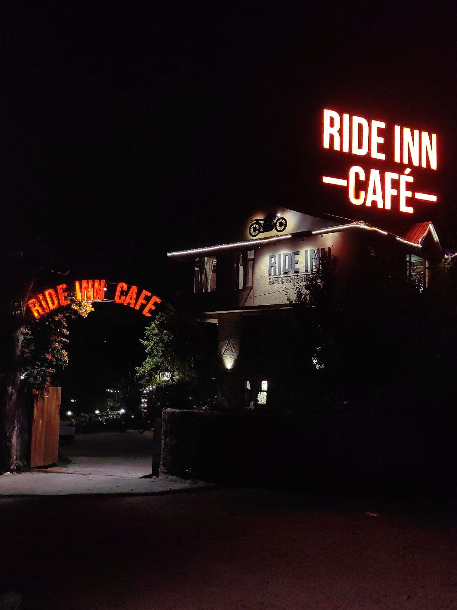 At night in Barheta, India, a neon sign with red lettering reads "RIDE INN -CAFÉ-" above a building. Another neon sign with red lettering, also reading "RIDE INN CAFE," is arched over an entryway to the left. The building has a dark roof with a red peak and is lit by string lights along the eaves. A silhouette of a motorcycle is visible on the roofline. Trees and shrubbery are present in the foreground and around the building, some adorned with string lights. The entryway is flanked by stone pillars and a wooden gate on the left. The ground in front of the entryway is a dirt or gravel path.