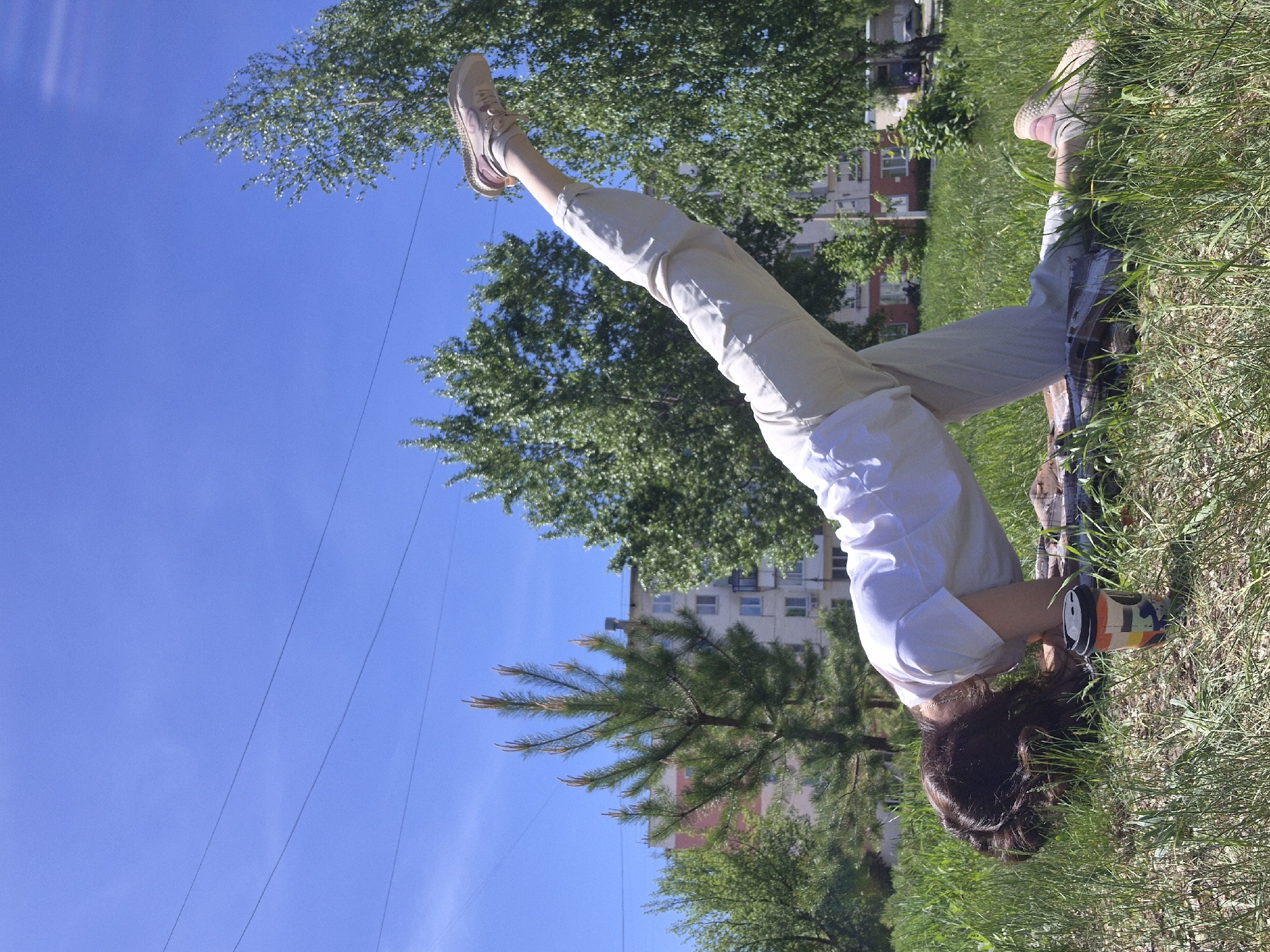 The image shows a person performing a headstand on a grassy area in an urban environment. The person is wearing a white shirt, light-colored pants, and sneakers. Their legs are extended upwards towards a blue sky with trees in the background. A building is partially visible in the background. The person's head is resting on a mat or blanket in the grass.