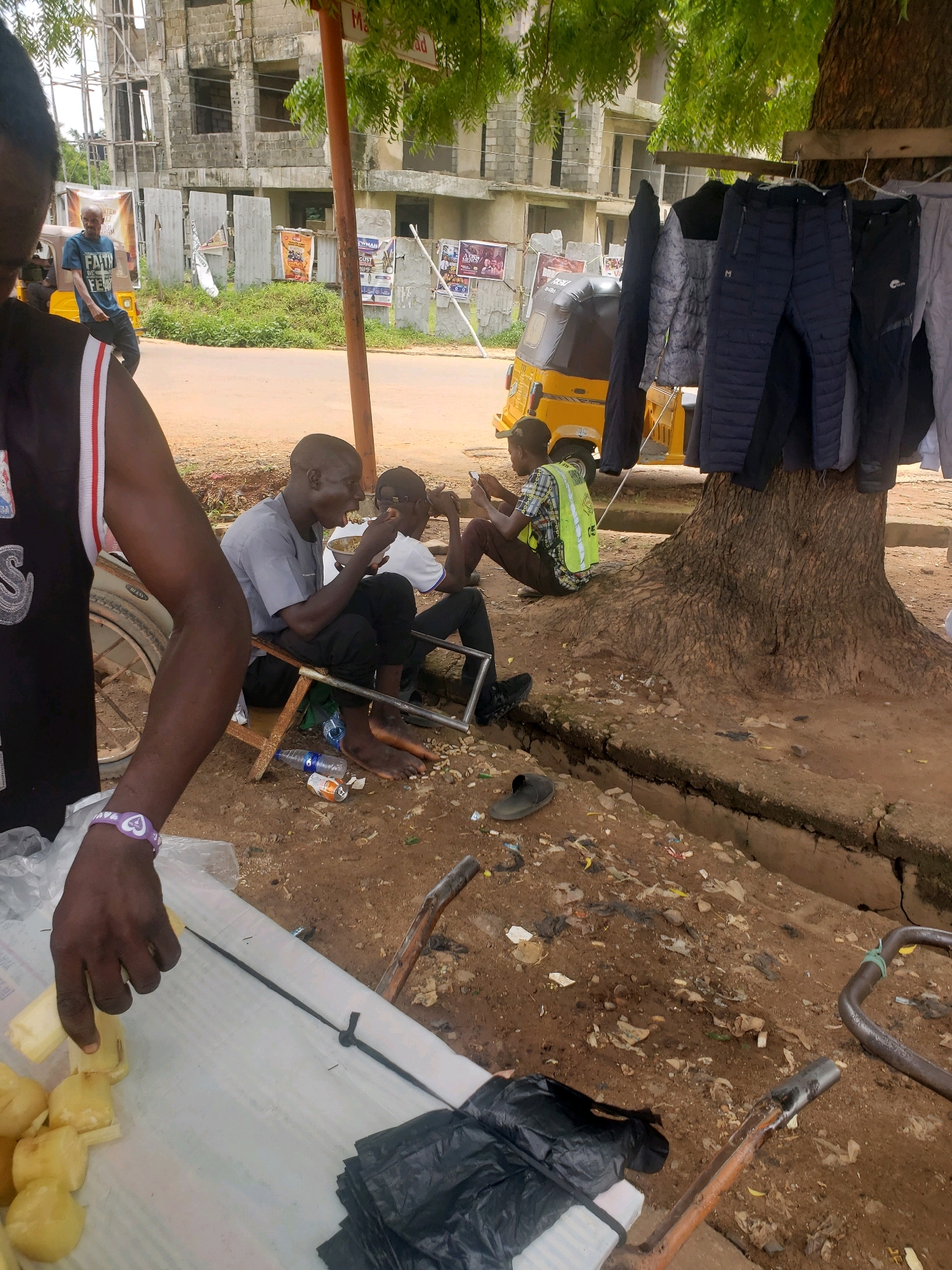 In Narayi, Nigeria, outside a construction site, two men sit on a metal bench. The man on the left, wearing a gray t-shirt, eats from a bowl with a spoon. The man on the right, wearing a yellow vest over a patterned shirt, looks at his phone. In front of them, a yellow auto-rickshaw is parked. Clothes, including jackets and pants, are hanging from a tree. In the foreground, a person's arm, wearing a purple bracelet, reaches for cut pieces of food on a white surface. The ground is covered in dirt, debris, and a cracked concrete channel. A partially constructed building is visible in the background.