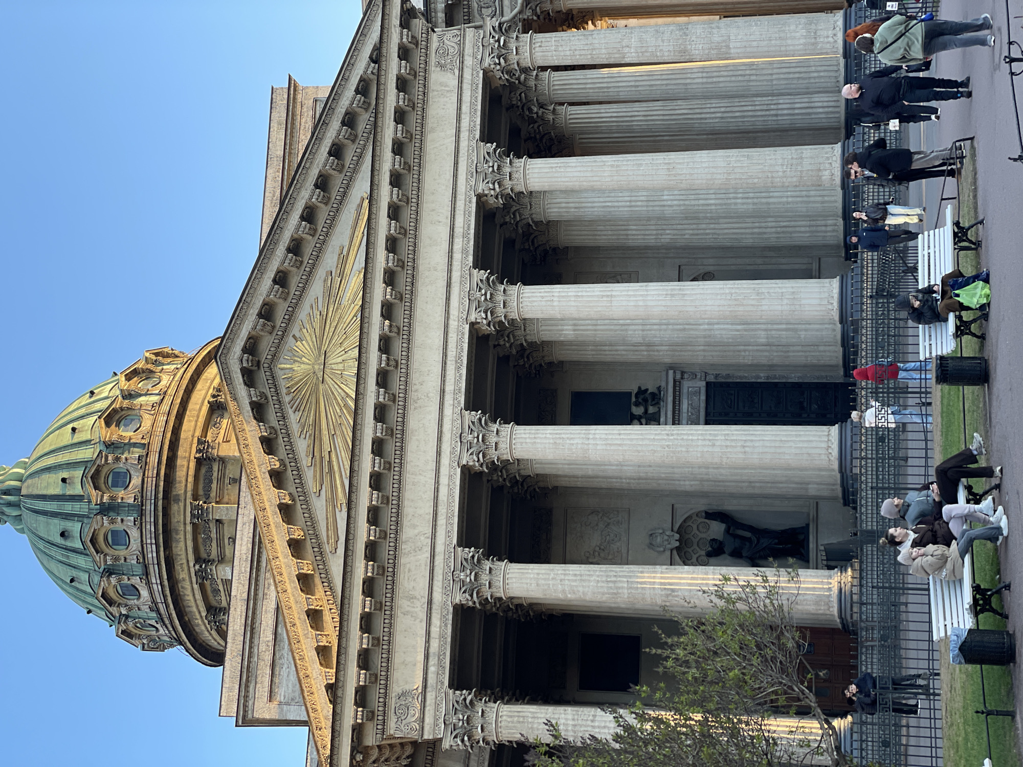 The image depicts the Kazan Cathedral in Saint Petersburg, Russia. A large dome with a green patina is partially visible on the left side of the frame, rising above a neoclassical facade. The facade features numerous tall, white columns with ornate capitals. A triangular pediment with a golden sunburst design is at the top of the structure.
People are visible in front of the building. Some are sitting on benches, while others are walking by. A black fence runs along the front of the building. There is also a tree with budding green leaves in the foreground on the bottom right corner. The sky is clear and blue.