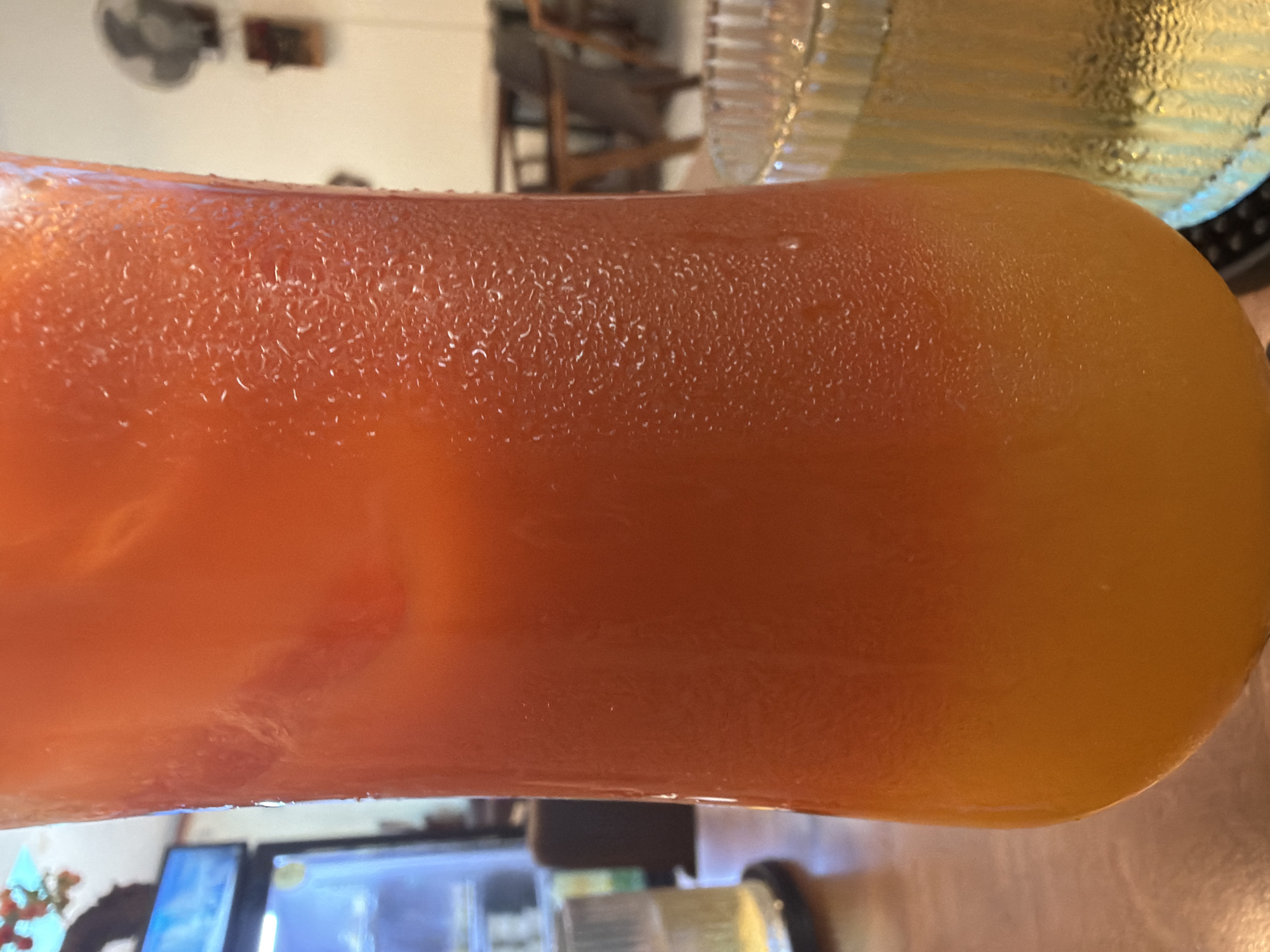 A close-up, eye-level shot focuses on a tall glass filled with a vibrant orange beverage, likely a juice or cocktail. The glass is covered in condensation, indicating it is cold. The beverage appears to have two distinct layers, with a darker orange at the top fading to a lighter, more yellow-orange at the bottom. Ice cubes are visible within the drink.
To the left of the main glass, a portion of another glass with a clear, pale yellow liquid is visible. This glass has a ridged texture. In the blurred background, a refrigerator with a blue display light is partially visible. Further back, there are tables and chairs, suggesting a dining or seating area, and a ceiling fan is mounted on the wall. The setting appears to be indoors, possibly a cafe or restaurant. The image's metadata indicates it was taken in the city of Bac Nha Trang Ward, Vietnam. There are no people clearly visible in the image.