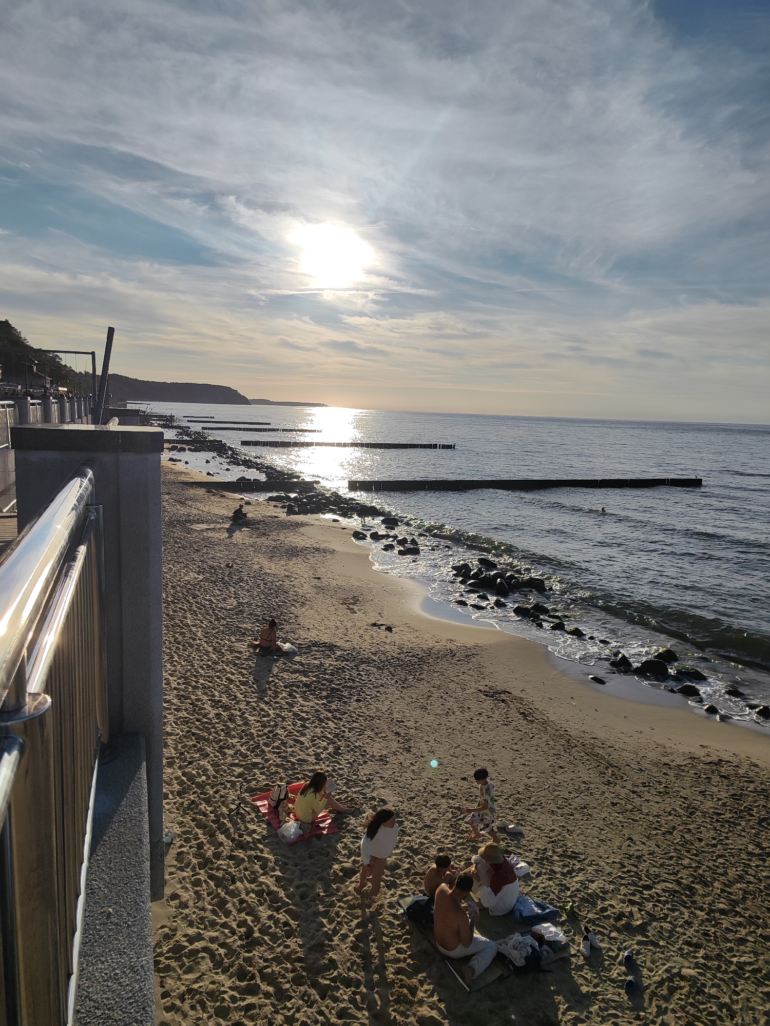 This image depicts a view of the Baltic Sea coast in Svetlogorsk, Russia, likely during late afternoon or early evening. The sun is setting or rising, casting a warm glow and reflecting off the water. The scene includes a sandy beach with several groups of people enjoying the coastline.
In the foreground, to the left, a concrete walkway with a polished metal railing runs along the edge of the beach. The walkway overlooks the beach and the sea. The beach itself is populated with individuals and small groups. One group, near the center of the image, consists of at least three people sitting on a red blanket, with one individual appearing to be eating. Further down the beach, another group of three to four people are gathered, some sitting on the sand. A child is visible near this group. Scattered across the sand are personal belongings such as bags and towels.
The sea stretches out to the horizon, with calm water and the sun’s reflection creating a bright path across its surface. Wooden breakwaters are strategically placed in the water, running perpendicular to the shore. The coastline curves in the distance, with a treeline and buildings visible along the elevated ground. Rocks are present along the shoreline, with some waves gently lapping against them. The sky is filled with wispy clouds, with the sun partially obscured by some of these formations. The overall atmosphere suggests a peaceful leisure activity by the sea.