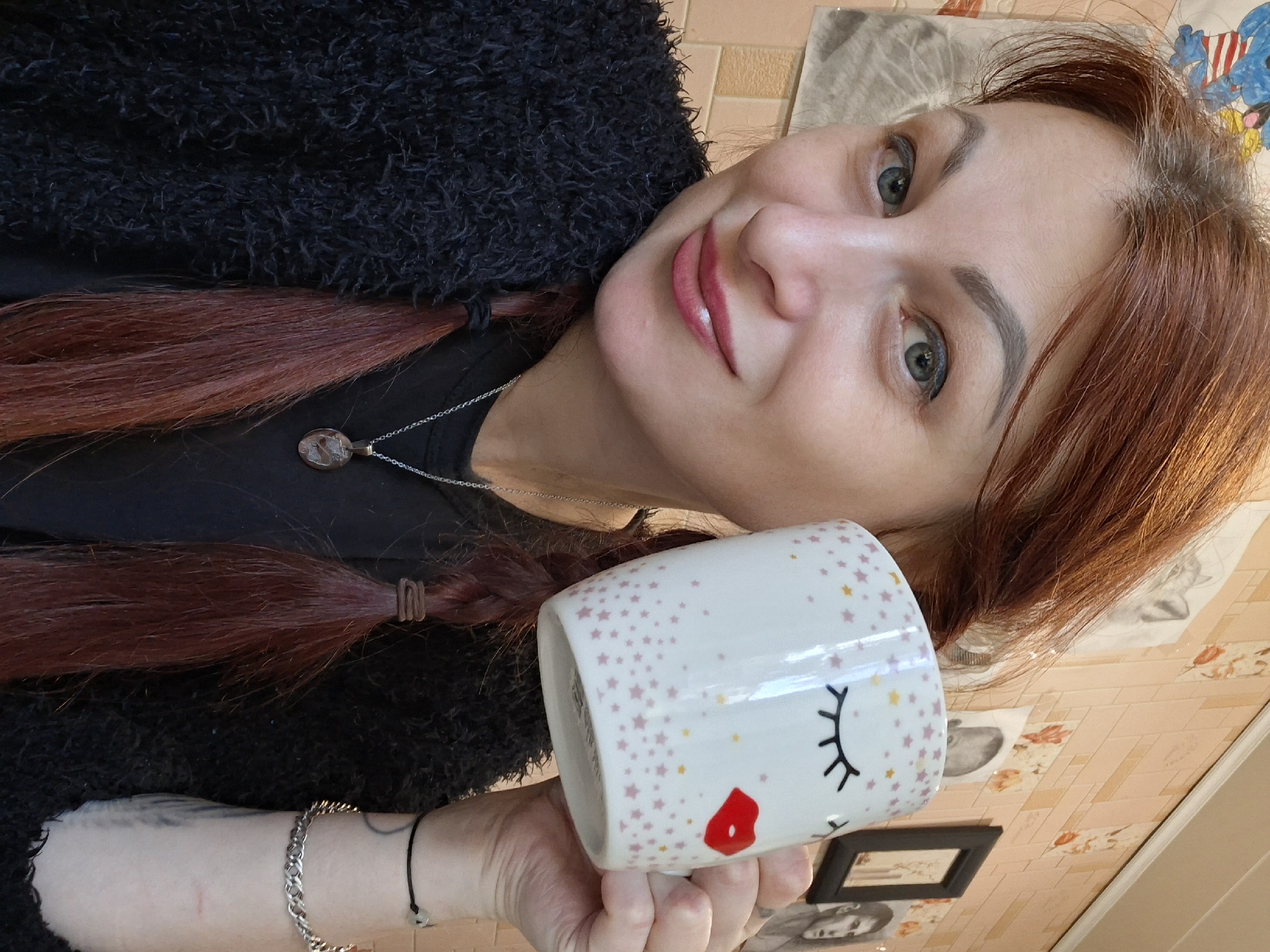 The image is a medium shot of a woman with reddish-brown hair styled in two braids. She is wearing a black, fluffy jacket over a black t-shirt and a silver necklace with a round pendant. She is holding a white mug decorated with colorful stars, closed eyes, and red lips up to her face, partially obscuring her right cheek. Her left hand is visible, also holding the mug, and she is wearing a bracelet on her right wrist. The background appears to be a wall with pink, textured tiles and several framed pictures and drawings. The lighting suggests an indoor setting. The subject is looking at the camera with a slight smile. The specific location in Myadzyel, Belarus is not determinable from the visual information provided.