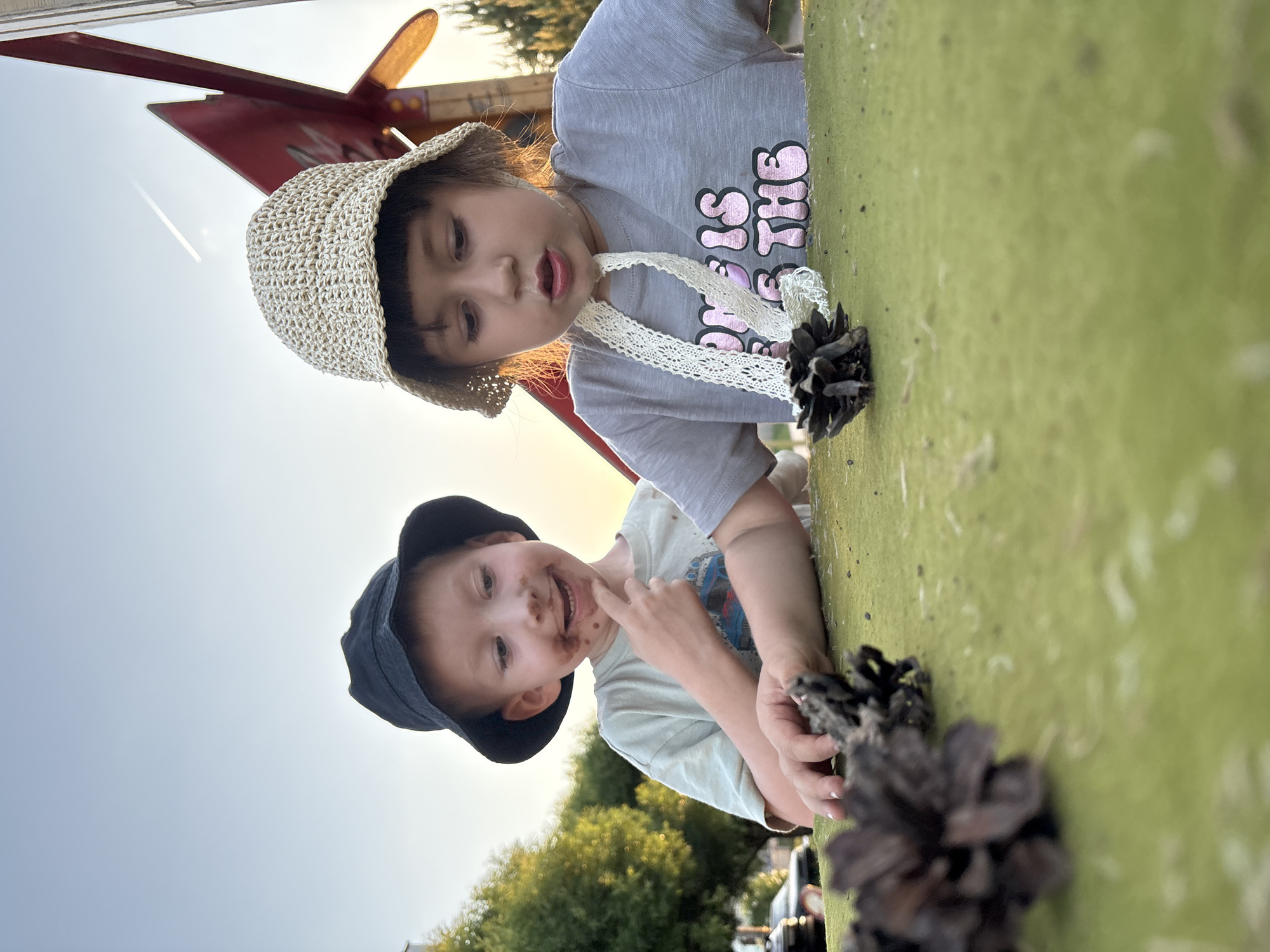 Two young children are leaning over the edge of a green surface, seemingly a structure within a playground or park. The child in the foreground has chocolate or dirt smeared around their mouth and is wearing a dark blue hat. The other child wears a woven hat and a gray shirt. Several pinecones are on the green surface. The presence of trees and parked cars suggests the location is an outdoor space, potentially a park, within Chaikovsky, Russia.