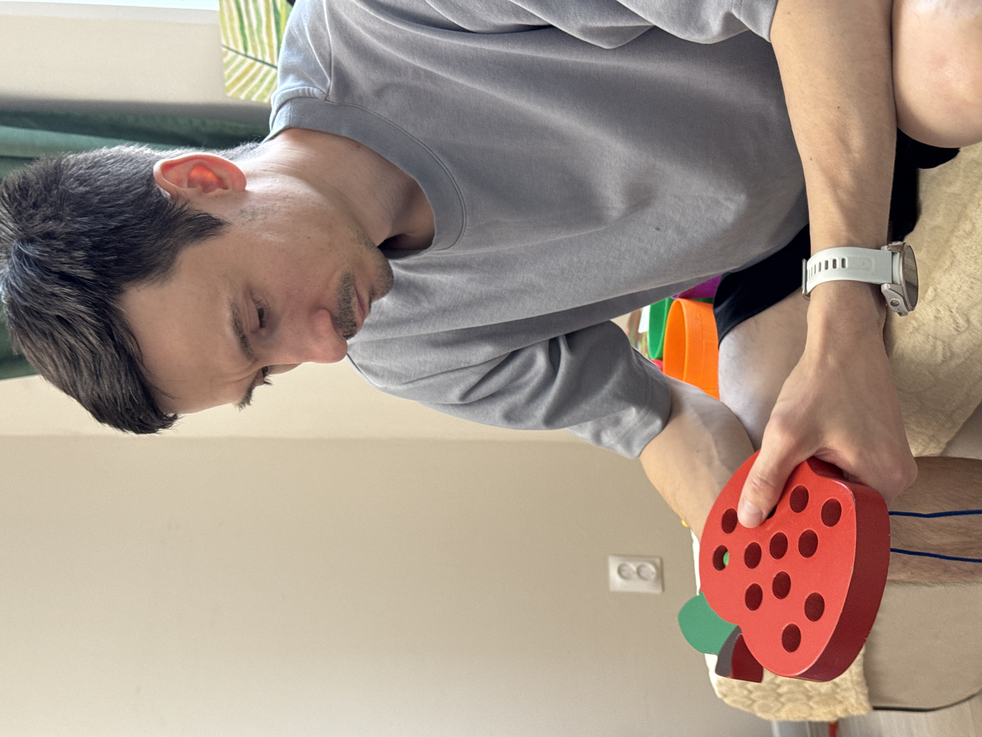 A male individual with short, dark hair and a mustache is depicted in a medium shot, looking downwards with a focused expression. He is wearing a plain, light grey, short-sleeved t-shirt and dark shorts. A white sports watch is visible on his left wrist. He is holding a red, wooden lacing toy shaped like a strawberry, which has multiple circular holes and a green stem, with a blue string hanging from it. He is seated on a light-colored, possibly upholstered surface with a textured, woven-looking throw draped over it. The background consists of a plain, light beige wall with a white double electrical outlet visible on the lower left. To the right, behind the individual, a dark green curtain, a portion of a window frame, and colorful plastic objects (orange, green) are partially visible. A minimalist piece of artwork with green striped patterns is also visible on the wall to the far right. The lighting is bright and even. The location, including the city of Izhevsk, Russia, cannot be determined from the visual information present in the image.
