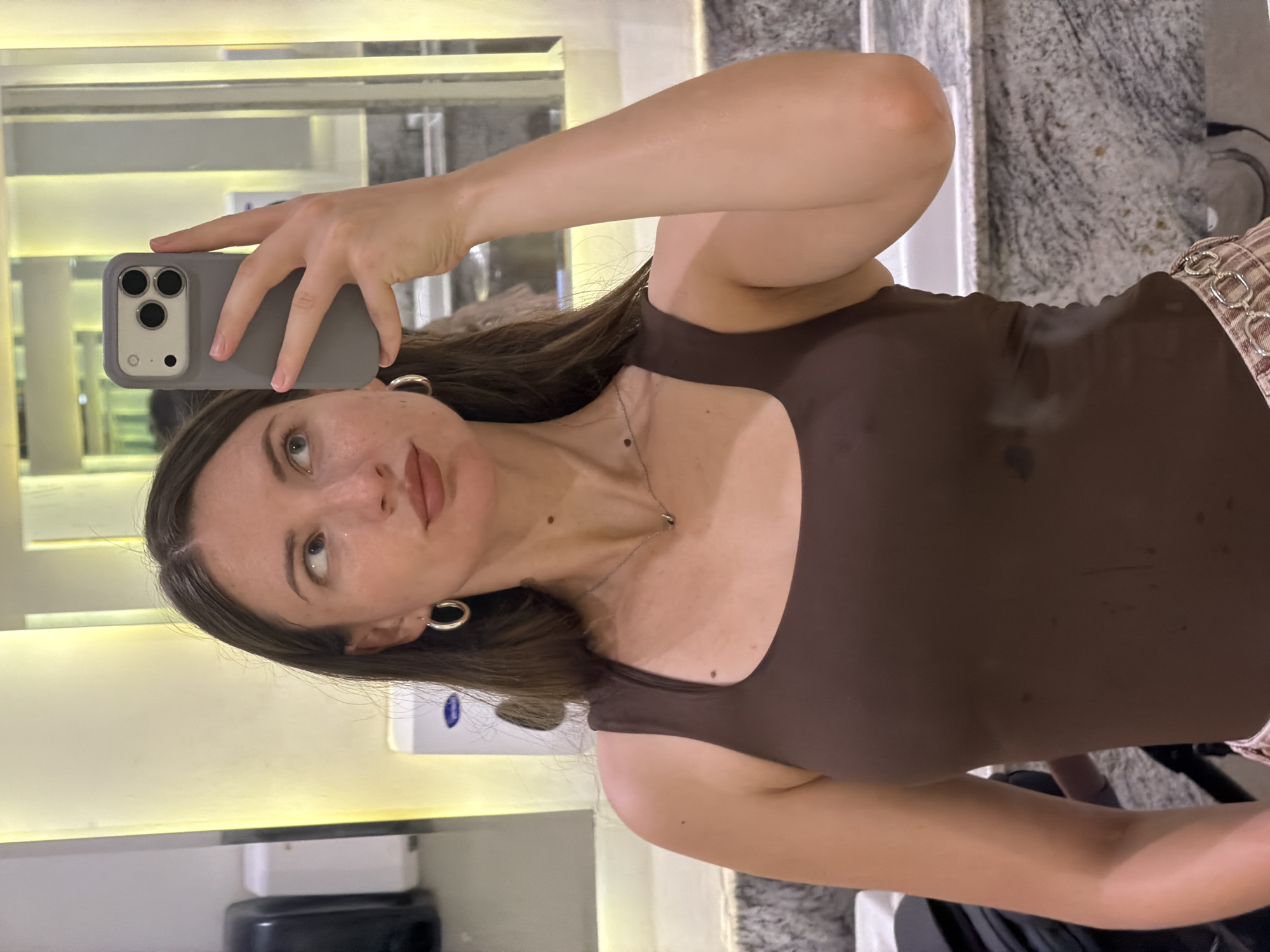 A young woman is taking a mirror selfie in a brightly lit restroom. She is wearing a brown tank top and a patterned skirt. The restroom appears to be in a hotel or resort in Sharm El Sheikh, Egypt, with marble countertops and illuminated mirrors. She is holding a gray smartphone in her right hand.