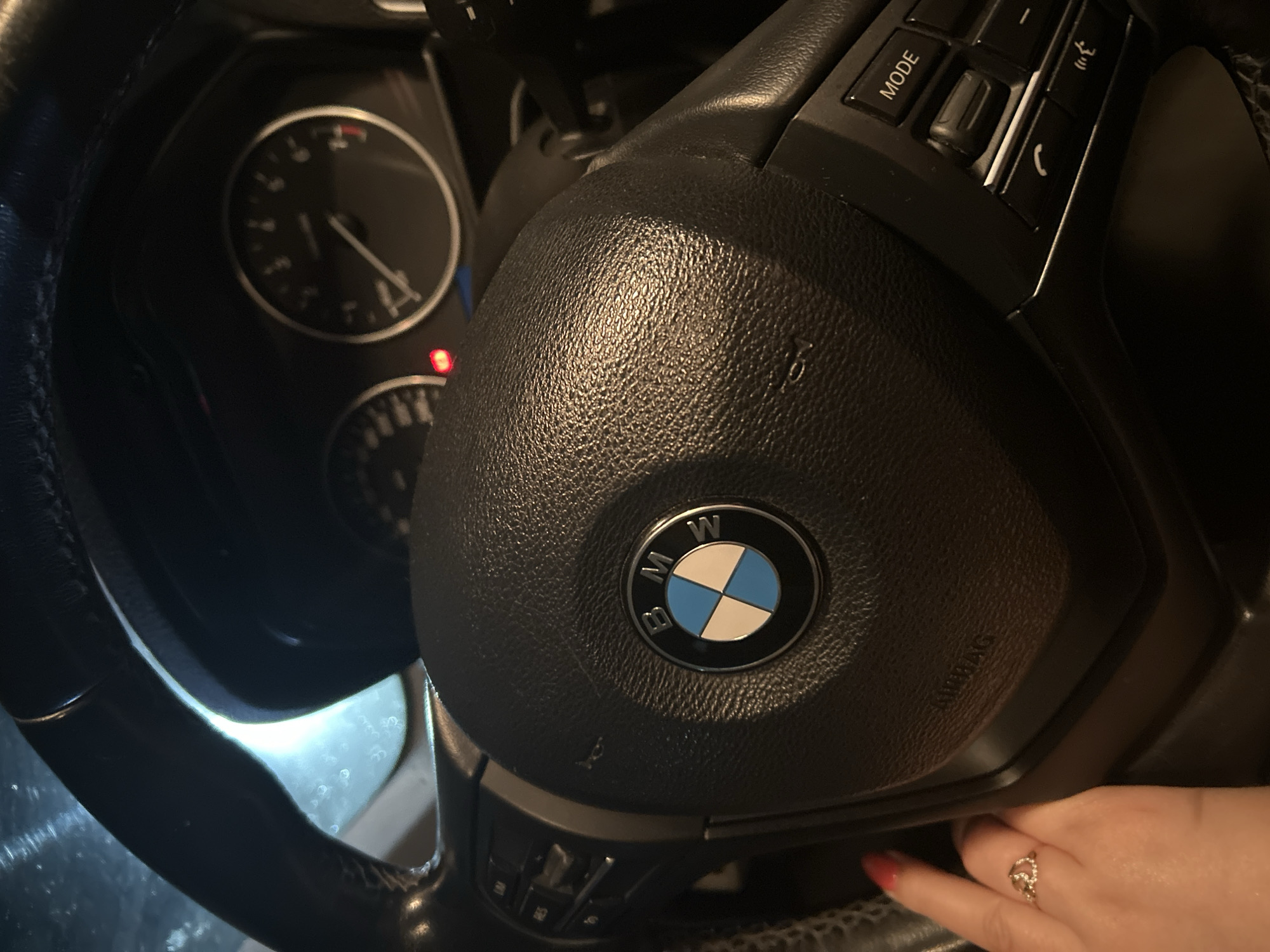The image shows a close-up view of a BMW steering wheel. The BMW logo is visible in the center of the steering wheel. To the left of the logo, there are buttons for cruise control and other functions. To the right of the logo, there are buttons for mode selection, volume control, and phone functions. The dashboard of the car, including the speedometer and tachometer, is visible in the background. A hand with red nail polish and a ring is resting on the steering wheel, on the left side of the image. The lighting suggests the photo was taken at night or in a dimly lit environment. The specific location in the city is unknown.