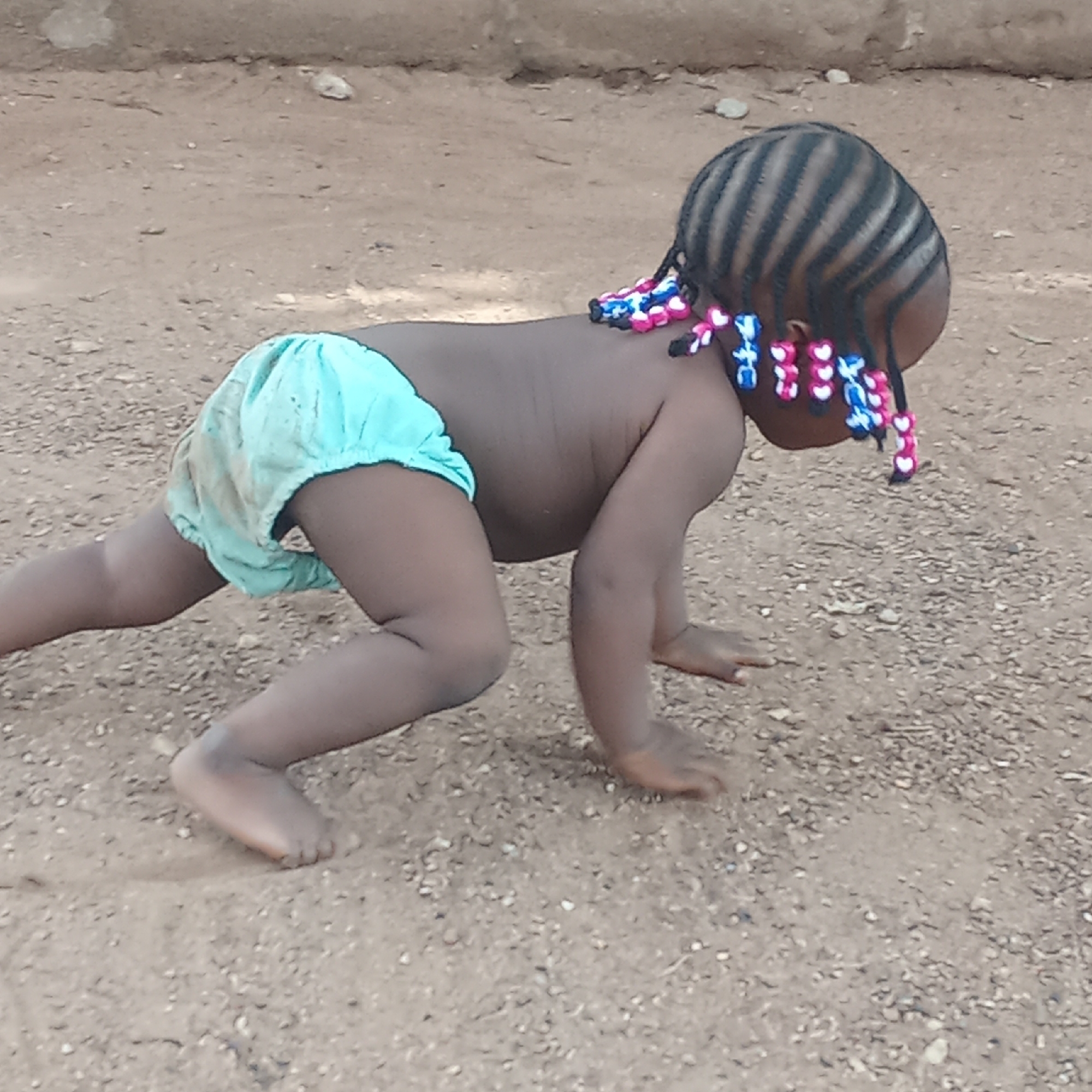 A young child crawls on all fours on a dirt surface. The child, who has dark skin, wears a light blue diaper. The child's hair is styled in cornrows with colorful beads at the ends. The background is a slightly out-of-focus wall. The location is Akwanga, Nigeria.