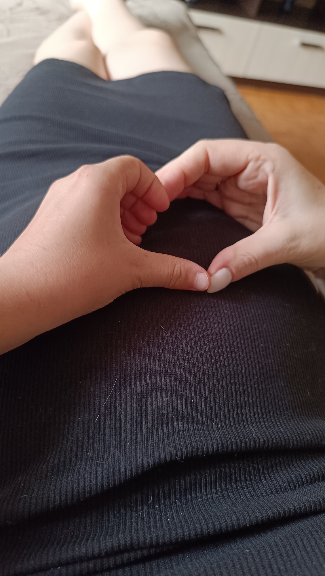 A person's hands are forming a heart shape over their lap, which is covered by a black ribbed garment. The background shows legs and a blurred piece of furniture. The scene appears to be indoors. There is no discernible connection to the city of Tolyatti, Russia.