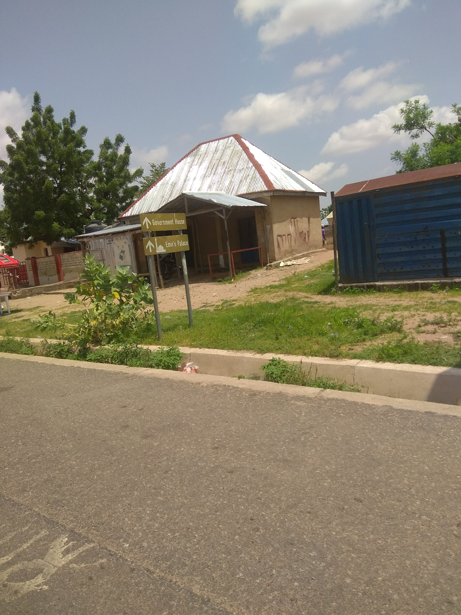 The image shows a street scene in Jalingo, Nigeria. In the foreground, a paved road curves to the left, with a concrete drain along its edge. A small, leafy green plant grows in the drain. In the middle ground, a signpost with two directional arrows stands on the right. The top arrow points to "Government House," and the bottom arrow points to "Emir's Palace." Behind the signpost, a small, light-brown building with a corrugated metal roof is visible. To the right of this building is a large, dark blue metal container. Further back, a large green tree with dense foliage stands on the left. In the background, more buildings and trees can be seen under a partly cloudy sky. A person in purple clothing is walking away from the camera in the distance. There are some scattered pieces of white litter in the drain.