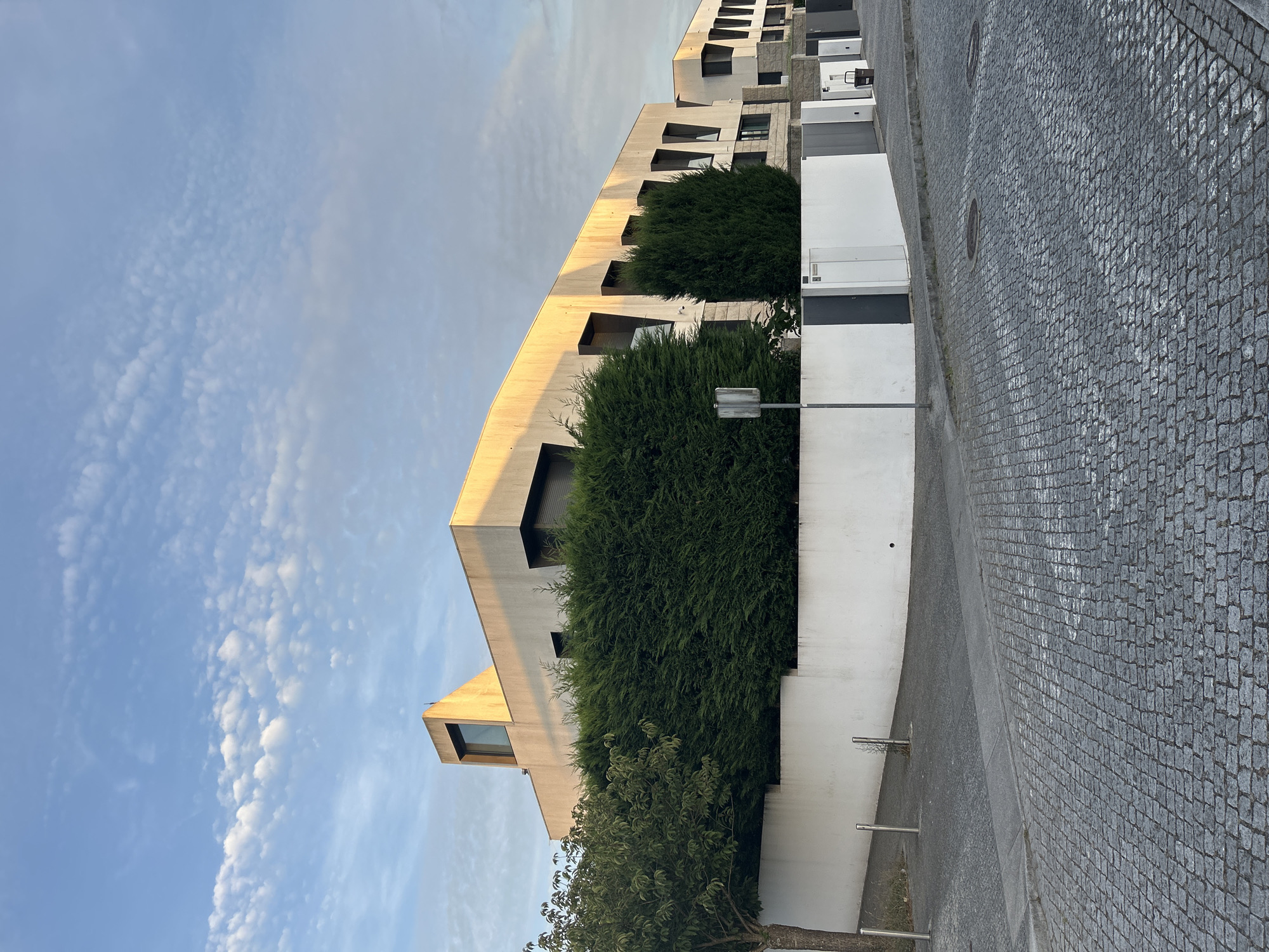 This is a photograph of a modern residential building in Porto, Portugal, captured during daylight. The building features a striking architectural design with a concrete facade and sharp, angular elements, particularly noticeable in the roofline and window frames. A prominent triangular window or skylight is visible on the upper section of the building. Lush green hedges line the front of the property, creating a strong visual barrier between the building and the street. A white wall runs along the perimeter, with sections of grey and white doors or gates integrated into it. The street in front of the building is paved with traditional cobblestones, typical of many European cities. The sky above is a mix of blue and white, with scattered clouds, suggesting fair weather. The overall impression is one of contemporary urban living with a touch of European charm.