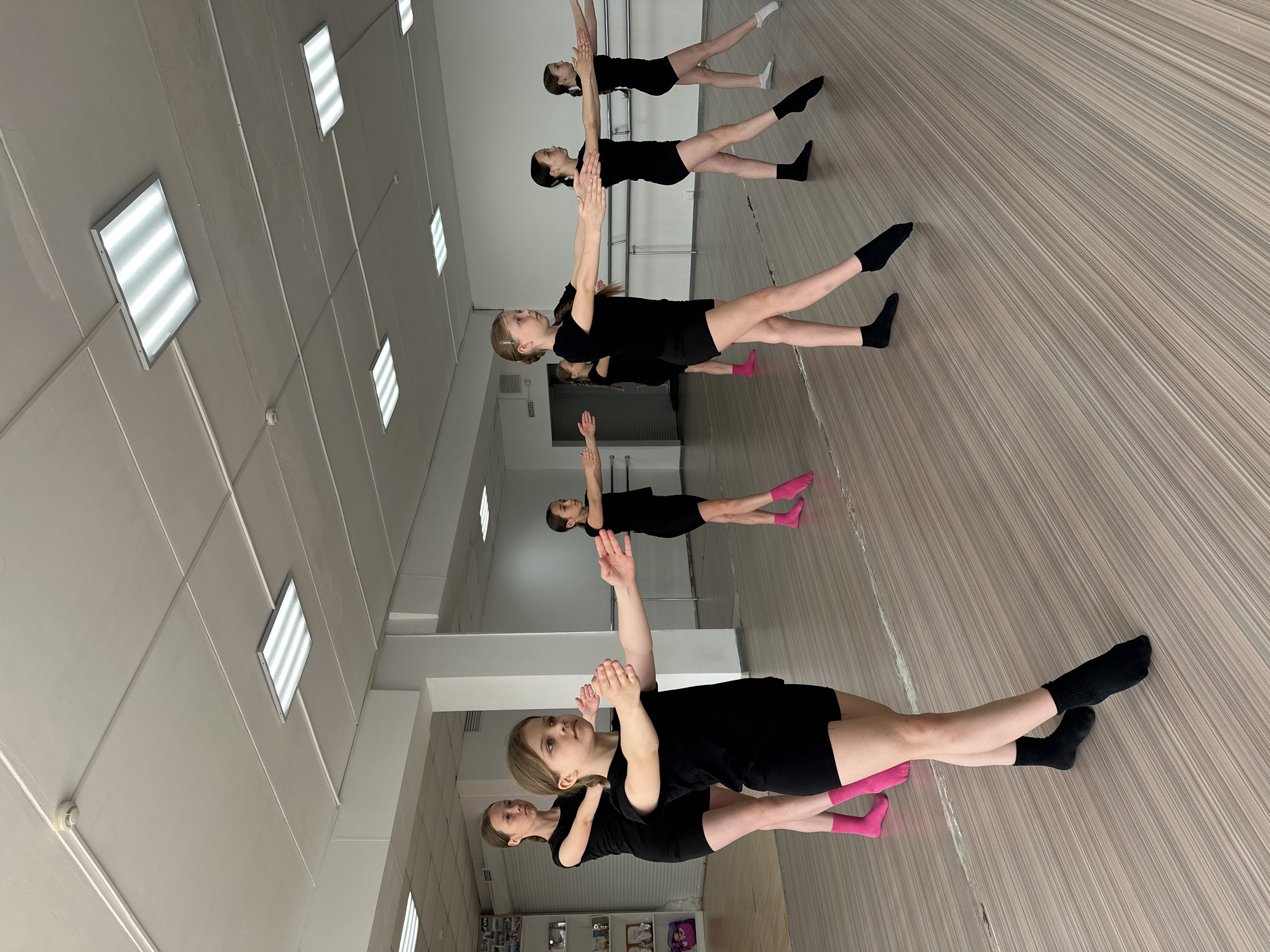 A group of young girls in black leotards and different colored socks (black, pink, white) are practicing a dance routine in a dance studio in Votkinsk, Russia. The girls are lined up in a row, with their arms raised and extended. The studio has a wooden floor, a white ceiling with rectangular light fixtures, and a mirror wall. The room has bright lighting and a clean appearance. A white wall can be seen behind the dancers with doors. A cabinet with personal belongings is against the wall on the lower left side of the image.