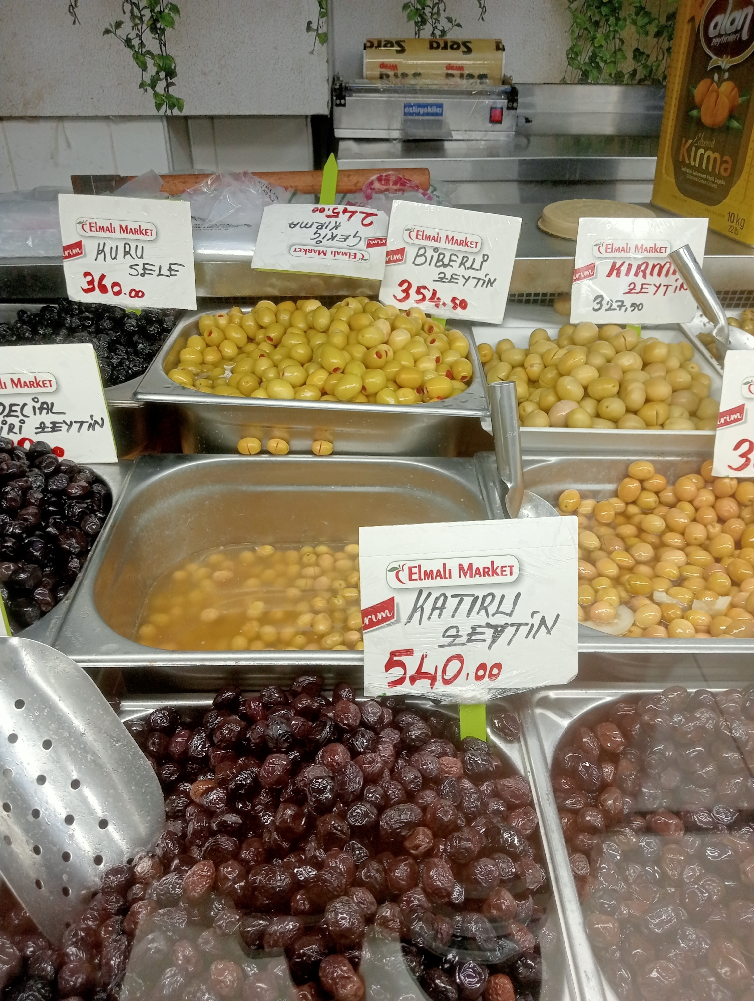 An eye-level, medium outdoor shot shows a market stall filled with olives. The stall is filled with various trays of olives. In the foreground, two large metal containers hold dark dried olives. A silver scoop sits in the left container. Behind these, two metal trays overflow with bright yellow-green olives. A third metal tray holds more yellow-green olives. In the background, a yellow bag of olives and a roll of plastic wrap are visible. White signs with black writing and red prices are placed in front of the olive trays. One sign reads "ELMALI MARKET" and "KURU SELE 360.00". Another sign says "ELMALI MARKET" and "BIBERLI ZEYTIN 354.50". A third sign states "ELMALI MARKET" and "KIRMA ZEYTIN 327.50". A fourth sign reads "ELMALI MARKET" and "KATIRLI ZEYTIN 540.00". The overall impression is one of a fresh produce market.