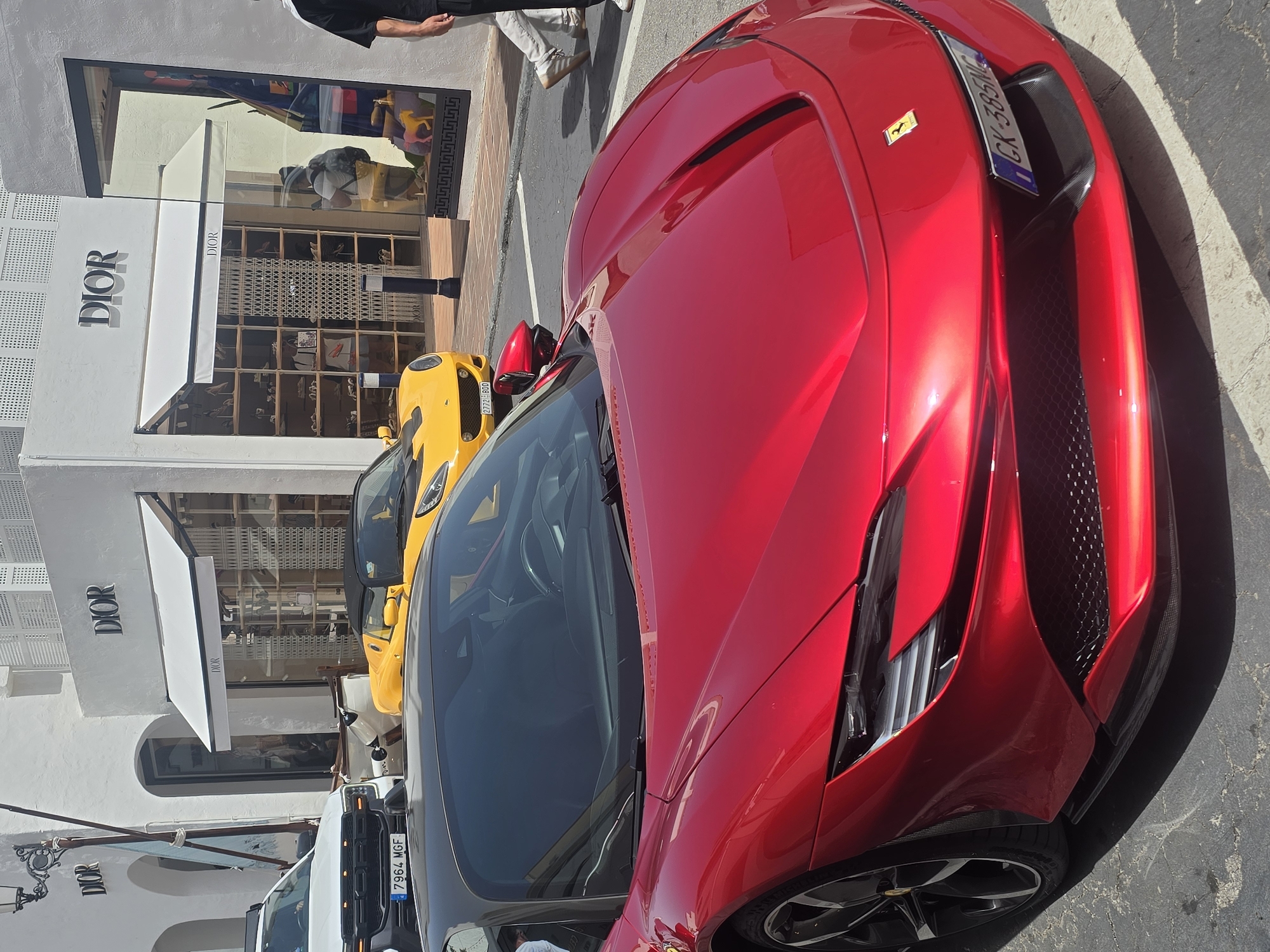A bright red Ferrari sports car is parked on a street in front of a white building with "DIOR" signage in Marbella, Spain. The Ferrari's hood and windshield are prominently displayed in the foreground. In the background, a yellow sports car is parked behind the Ferrari, and a white SUV is to the left. A person is walking away from the Dior store, which has a large display window showcasing various items. The street is paved, and there are white lines marking parking spaces. The architecture suggests a commercial area, likely a high-end shopping district.