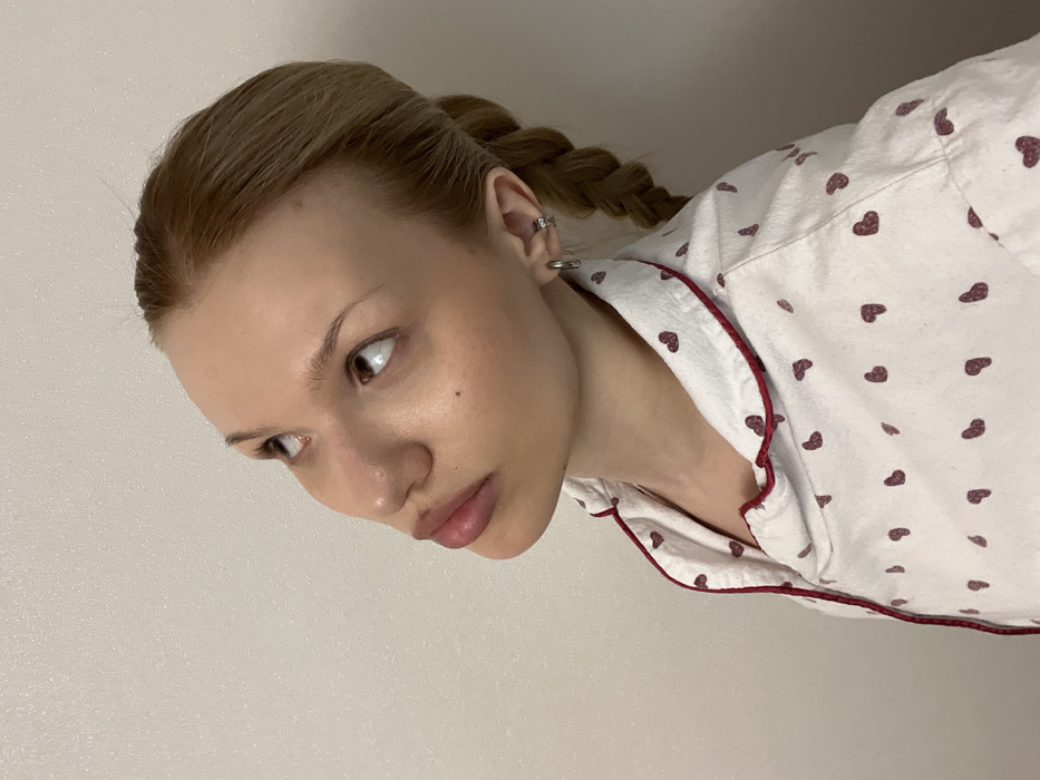 A young adult female is depicted in a close-up, chest-up shot against a plain, light beige textured wall, presumably indoors in the City of Chaikovsky, Russia. Her head is tilted upwards and slightly to the right, with her gaze directed upward and to the left. She has light brown hair styled in a visible braid on the right side of her head. She is wearing a white collared shirt patterned with numerous small maroon hearts and edged with maroon trim. A silver-colored hoop earring is present in her right earlobe, and a small silver-colored cuff earring is on the helix of her right ear.
