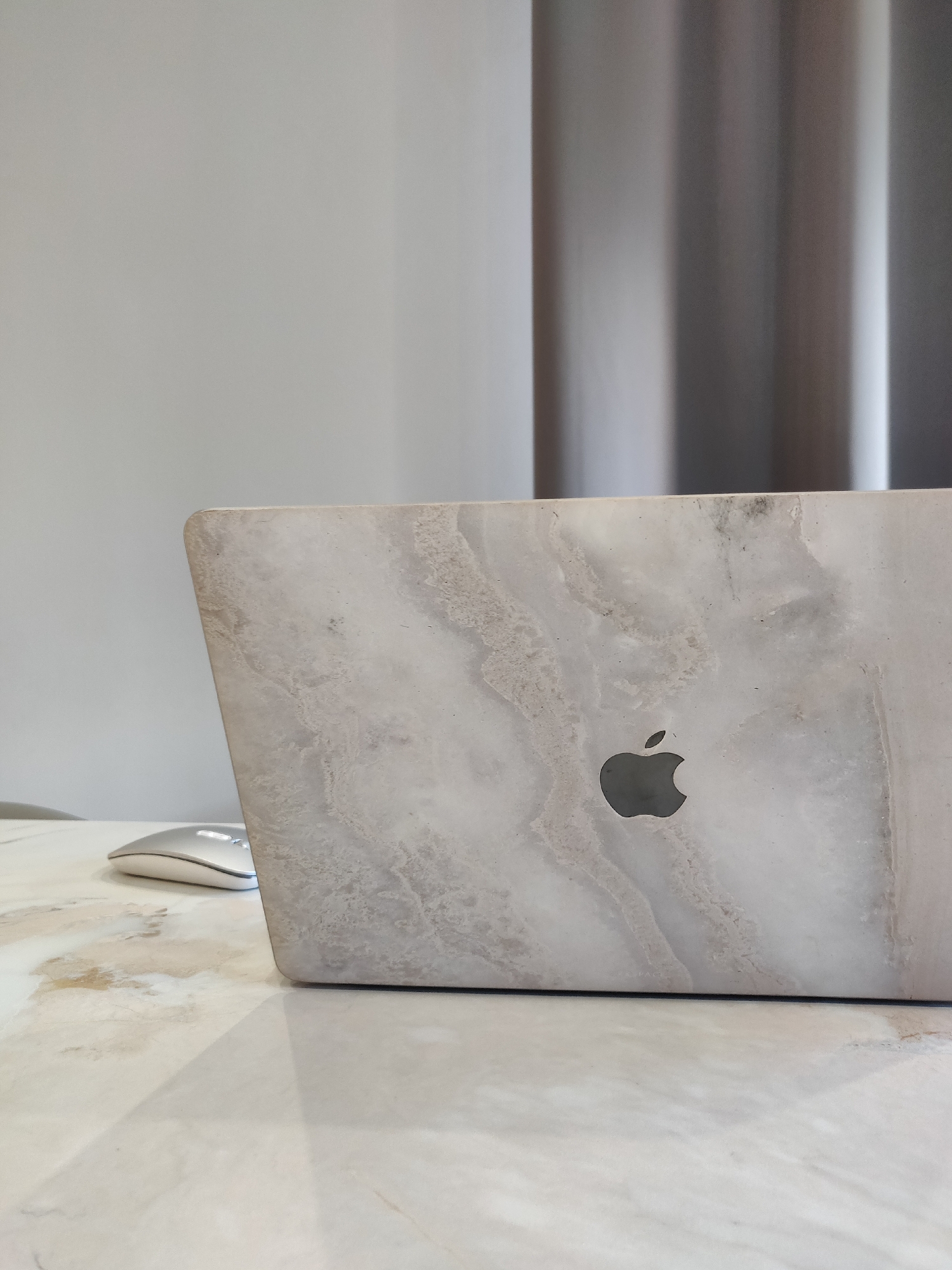 A laptop with a faux marble skin is partially visible on a matching faux marble table. A silver wireless mouse is on the table near the laptop. A grey wall and curtains are in the background. The image does not provide enough information to determine a location in Chaikovsky, Russia.