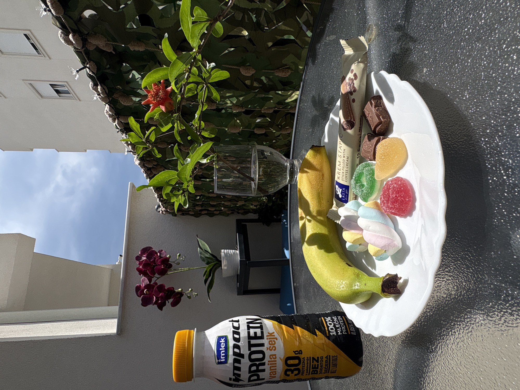 The image shows a table setup outdoors, likely on a balcony or terrace, given the presence of buildings and a glimpse of the sky in the background. A white bottle of "Imlek Impact Protein" vanilla shake with "30g" printed on the label sits near the bottom left. A white plate is positioned on the right side of the table, holding a banana, a "Lasta Krem Stapic" stick, chocolate pieces, jelly candies, and marshmallows. A glass and a flowering plant in a glass vase are placed near the plate. The background shows more greenery, parts of buildings, and the sky. The scene seems to be in a sunny environment, probably in Bar, Montenegro.