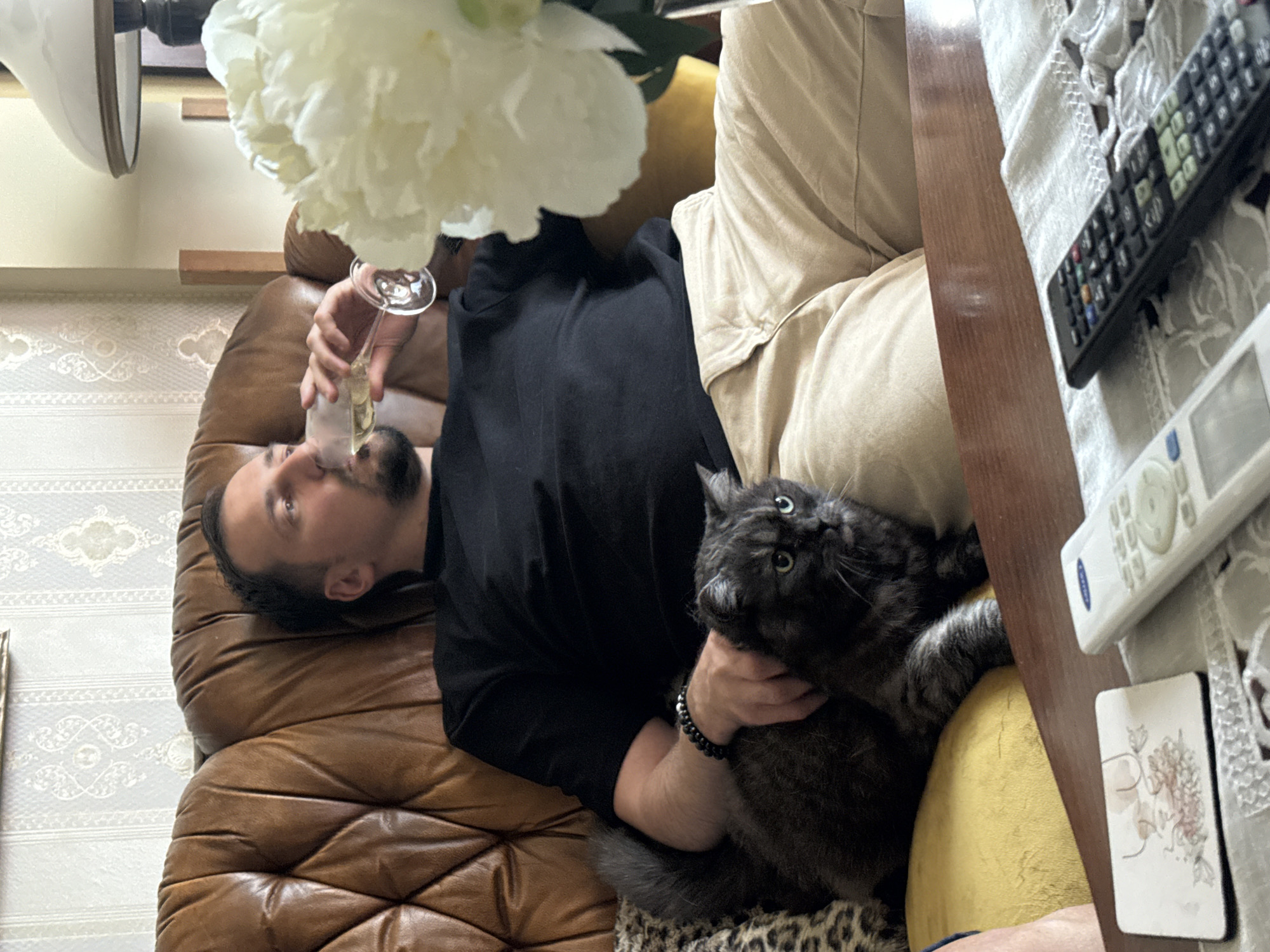 A man is lying on a brown leather couch, holding a champagne glass to his lips, appearing to drink. He is wearing a black shirt and has a beard. A grey cat is resting on his chest. A bouquet of white flowers is positioned above the man. On a table adjacent to the couch, there are two remote controls. The location is not specified.