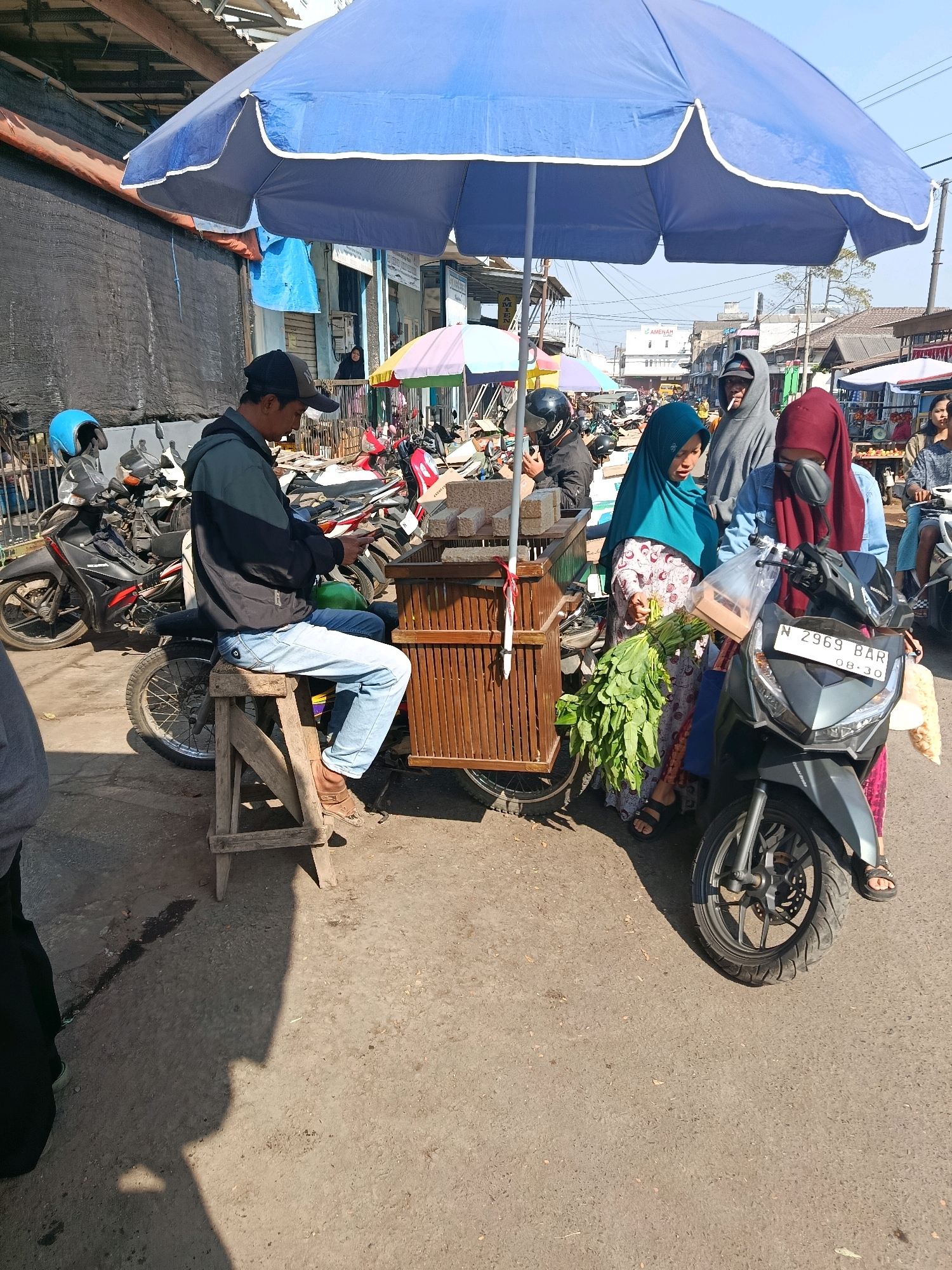 In Bululawang, Indonesia, a market scene unfolds under a large blue umbrella. A man sits on a wooden stool beside a cart filled with goods, appearing to be engaged with his phone. Two women stand near a motorcycle, one holding a bundle of leafy green vegetables. The environment is outdoors, with other market stalls, motorcycles, and buildings visible in the background, suggesting a bustling street market. The day is bright, casting long shadows on the ground.