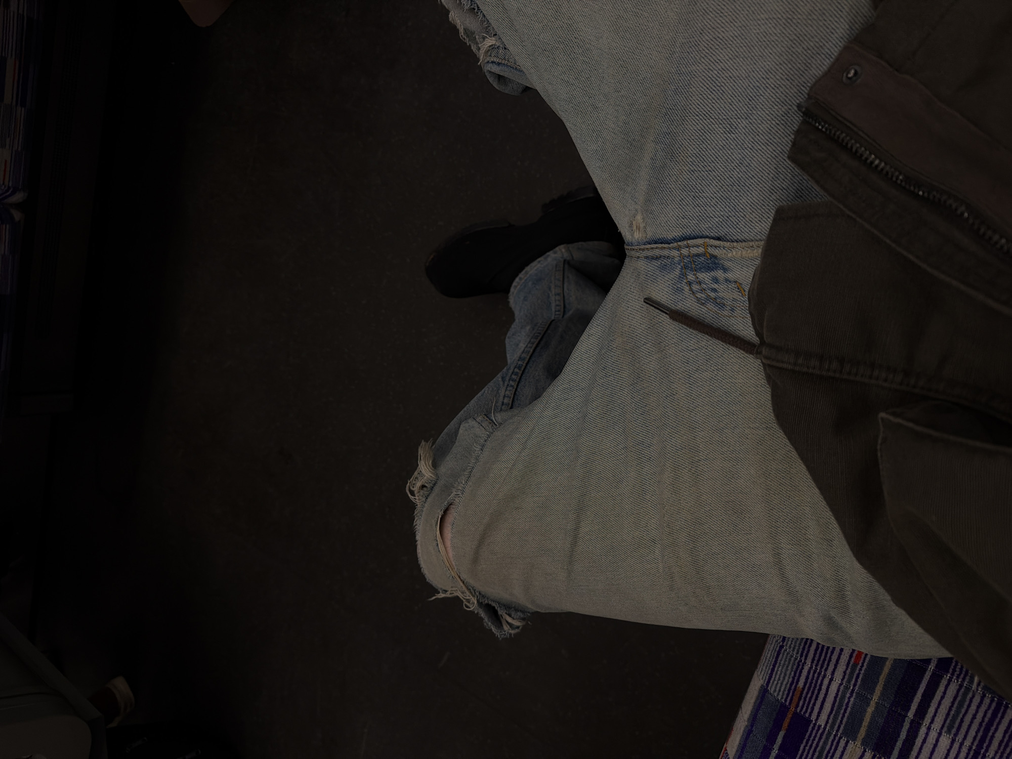 The image presents a ground-level perspective of a person seated, likely indoors given the controlled lighting. The individual is wearing distressed light-wash denim jeans, featuring a significant rip at the left knee. A black shoe is visible on the left foot, positioned on what appears to be a dark, possibly black, flooring. A dark-colored jacket rests on the right side of the person's lap, with a visible zipper and pocket detail. A silver zipper pull extends towards the jean-clad leg. A section of patterned fabric is visible underneath the jacket, suggesting the seat's upholstery. The setting is indeterminate based on the visual information provided. There is no specific information available to deduce the location as London, United Kingdom.