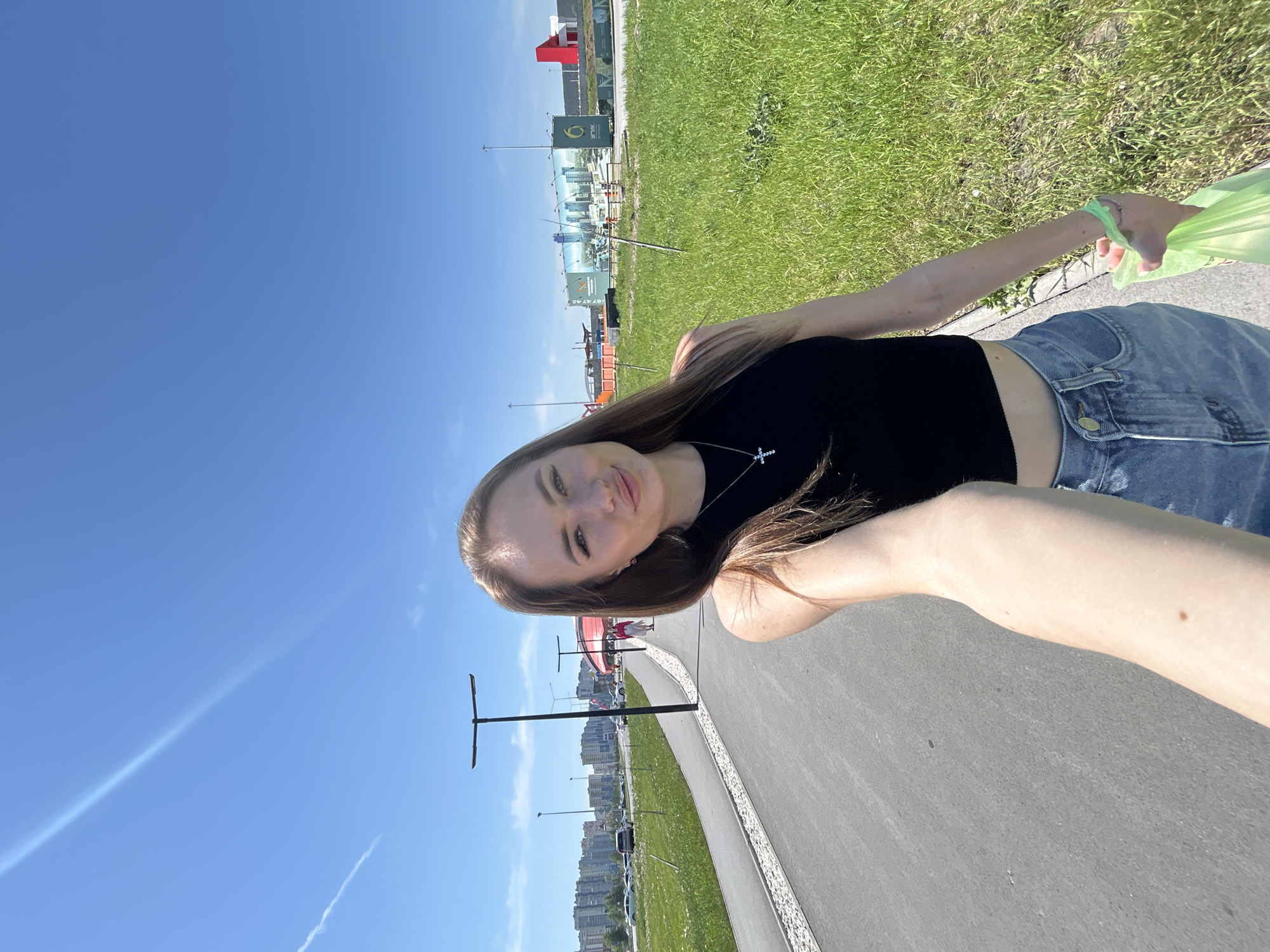 A young woman is depicted taking a selfie outdoors on a bright, sunny day. She has long, light brown hair, wears a black sleeveless crop top, light blue denim jeans, and a silver cross necklace. Her right arm, extended towards the camera, shows a distinct mole on the forearm. She holds a translucent light green item in her left hand.
The setting features a dark asphalt pathway, bordered on the right by white decorative stones, stretching into the background. Lush green grass and some wildflowers flank both sides of the path. Two modern black streetlights with horizontal fixtures are visible along the left side of the path.
In the far background to the left, a cluster of multi-story residential buildings forms a city skyline, with a road and several cars in front of them. To the right, large commercial billboards, one displaying a cityscape image and "УГМК" text, stand before industrial-style buildings. One of these buildings has a prominent red architectural feature. The sky is clear blue with a long, bright contrail extending diagonally across the upper left quadrant. Two indistinct human figures are visible further down the pathway in the mid-distance.