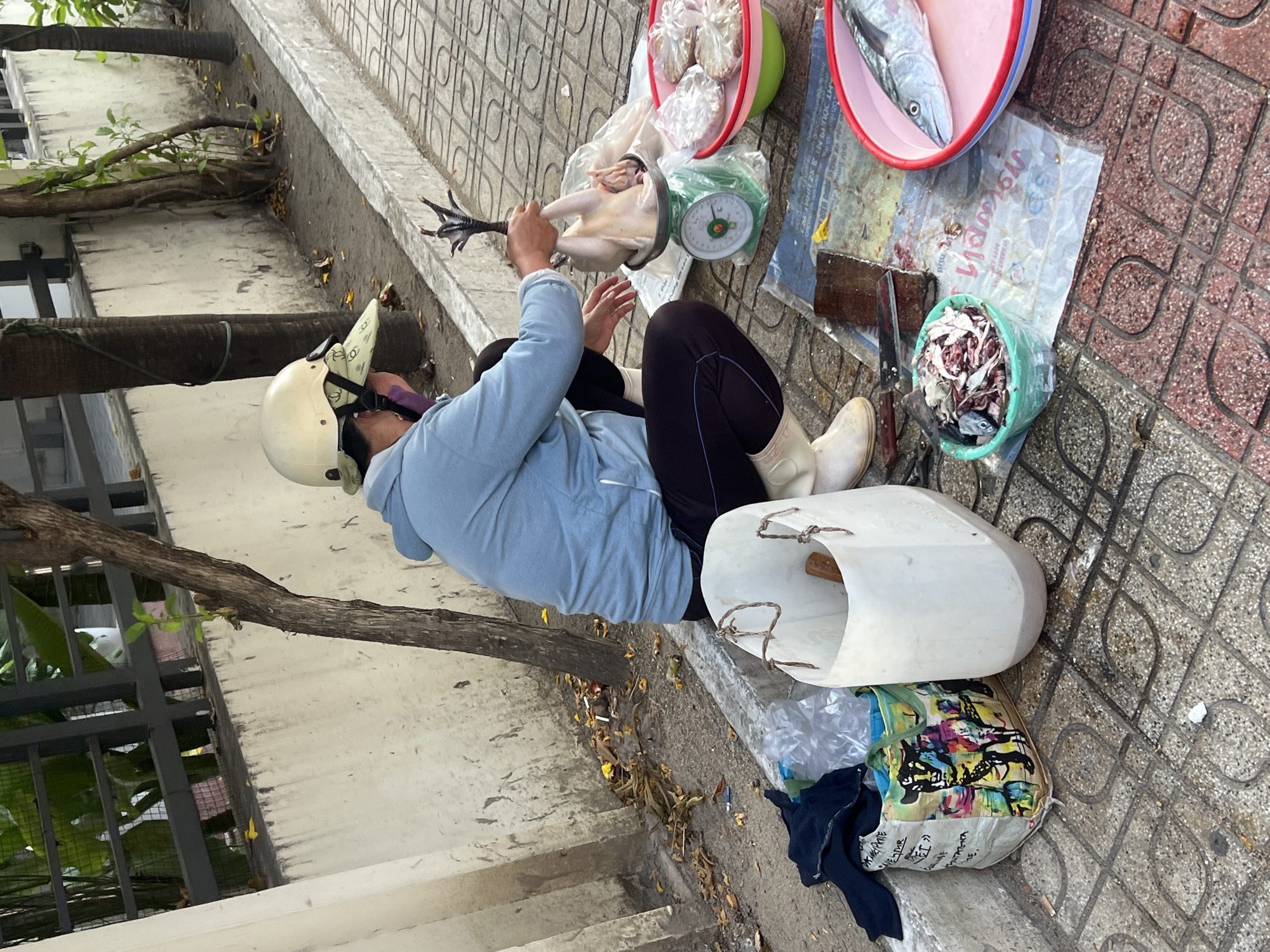 A woman wearing a light blue hooded shirt, dark pants, white boots, and a cream-colored helmet is sitting on the sidewalk. She is holding a whole chicken by its feet, placing it on a scale. Next to her are buckets containing fish, chicken meat, and individually bagged items. A cleaver rests on a cutting board near the buckets. The setting appears to be an outdoor market area. This scene is most likely located in the city of Nha Trang, Vietnam.