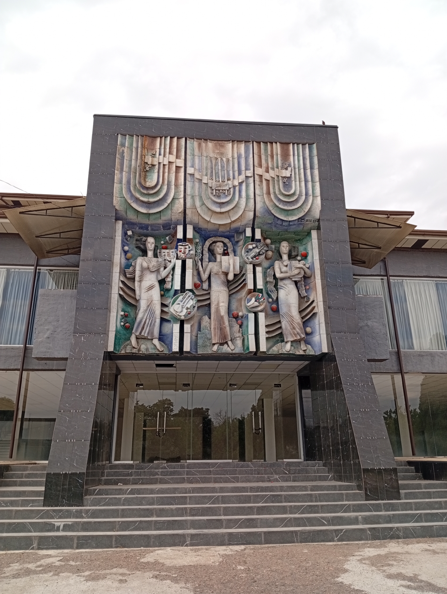 The image shows the facade of a building in the city of Angren, Uzbekistan. The building features a prominent, colorful relief sculpture above the entrance. The sculpture depicts three female figures in flowing robes, surrounded by abstract circular elements and smaller decorative objects. The figures appear to be holding or interacting with various symbolic items, such as masks and what might be musical instruments. The relief is mounted on a dark, textured surface, which is part of the building's facade. Below the relief is a set of glass doors, accessed by a wide, multi-tiered stone staircase. The staircase is made of dark, possibly granite, stone with lighter veining. The building itself has large glass windows on either side of the entrance, with sheer white curtains visible inside. Awnings extend from the upper part of the building. The sky above is overcast. The overall impression is of a public building, possibly a cultural institution or a government office, with artistic embellishments.