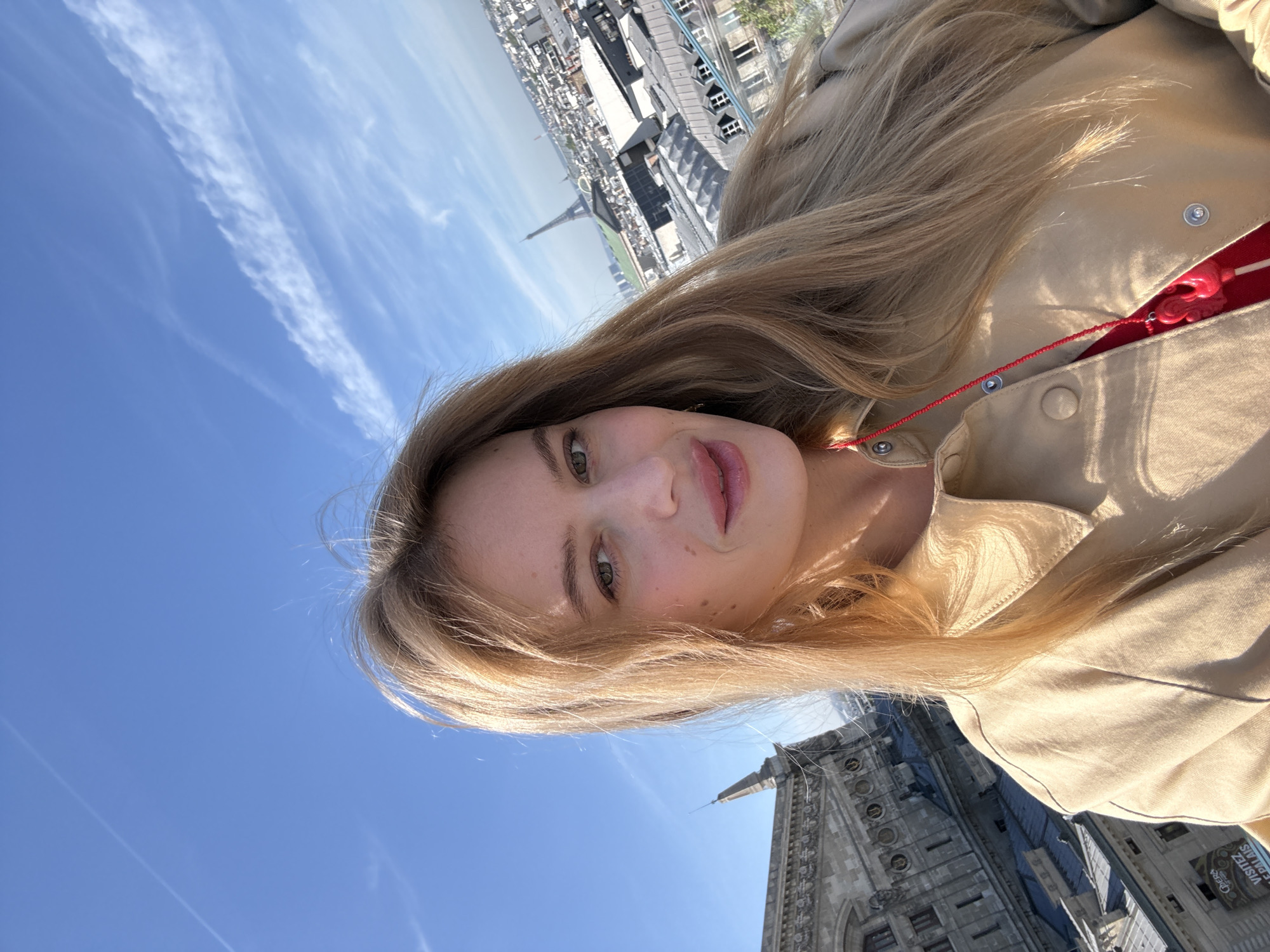 A woman with long blonde hair takes a selfie. In the background is the Eiffel Tower and a building. It is a sunny day with blue skies and scattered clouds. The picture appears to be taken from a high vantage point near a building with a spire.