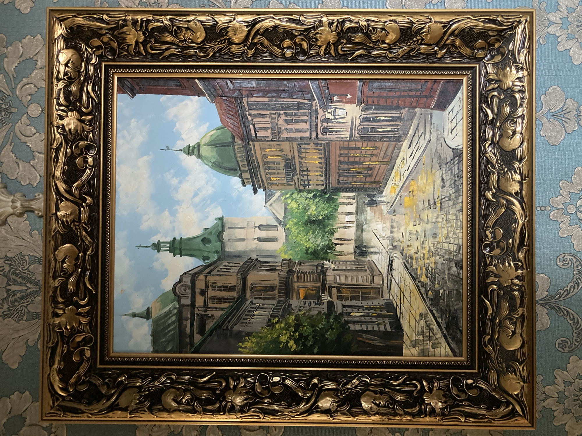 The image displays a framed painting of a European-style city street scene, possibly after a rain shower, given the reflections on the wet cobblestone. The painting is vertically oriented and depicts historical architecture with green domed roofs, suggesting a church or similar prominent building. Ornate buildings line the street, with intricate architectural details visible. The sky is a bright blue with fluffy white clouds, indicating daytime.
The painting itself is rendered in a realistic style, with noticeable brushstrokes that give it texture and depth. The play of light and shadow on the wet pavement and building facades is a prominent feature. The colors are rich, with warm tones in the buildings contrasting with the cool blues of the sky and the vibrant green of the domes and foliage.
The painting is enclosed in an elaborate, gilded frame with ornate carvings of floral and possibly animal motifs. The frame adds a sense of grandeur to the artwork. The painting is hung against a textured blue wallpaper with a subtle floral pattern.
No people are clearly discernible in the painting, though the scene appears to be a public space that would typically see pedestrian traffic. The overall mood of the painting is one of serene beauty and historical charm, perhaps evoking a sense of nostalgia or contemplation of the past. There is no visible text in the painting or the frame.