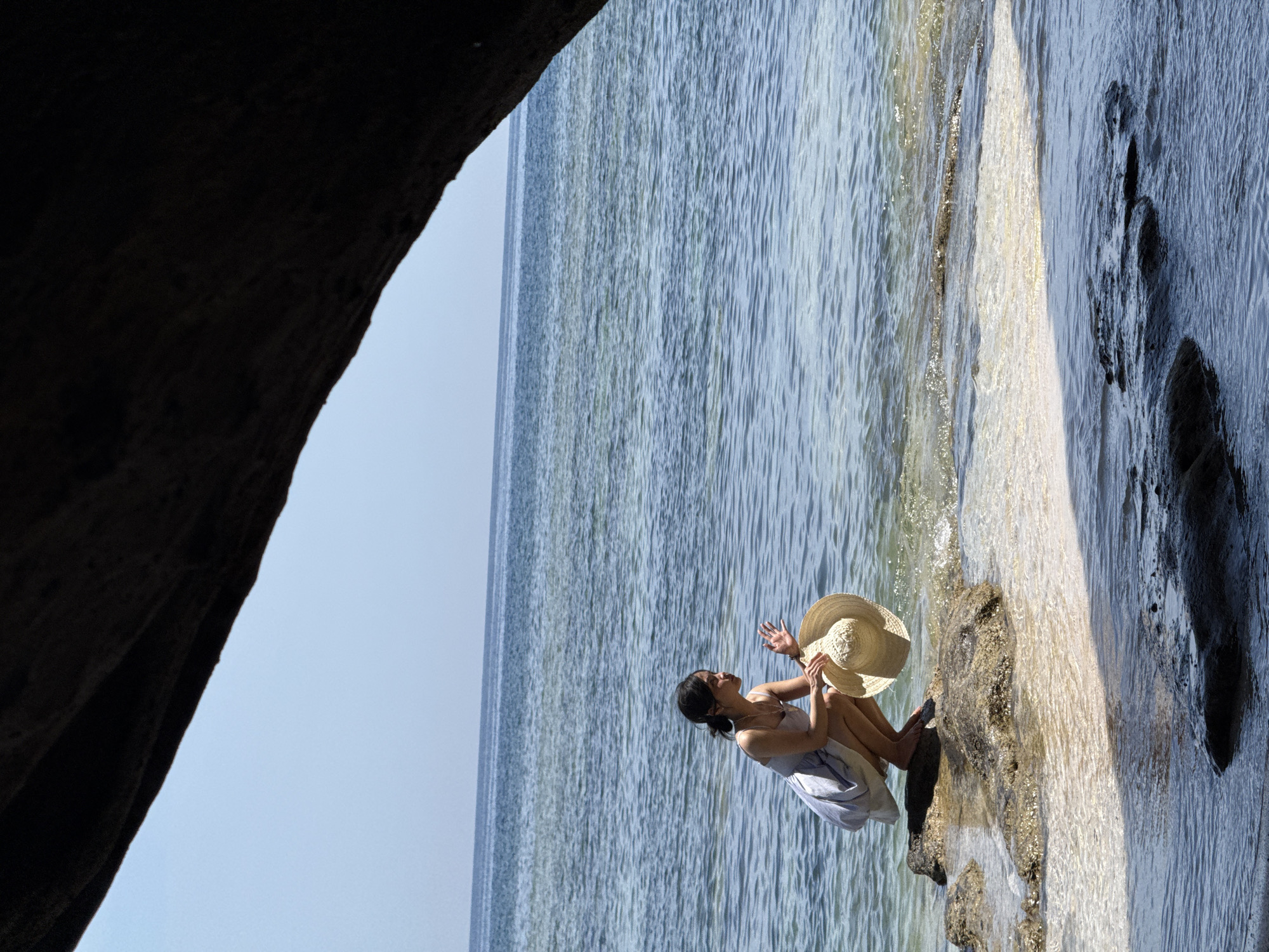 A young woman wearing a light-colored sundress is crouched on a rock at the edge of the water, holding a straw hat in her right hand. She is looking out at the ocean and raising her left hand as if to wave. The water is clear and blue, with gentle waves lapping at the shore. The scene is framed by a dark, rocky overhang, suggesting a cove or a sheltered spot on the coast. The sky is clear and blue, and the overall impression is one of a serene day at the beach. This appears to be a coastal area, possibly on the island of Lý Sơn, known for its volcanic landscapes and beautiful beaches.