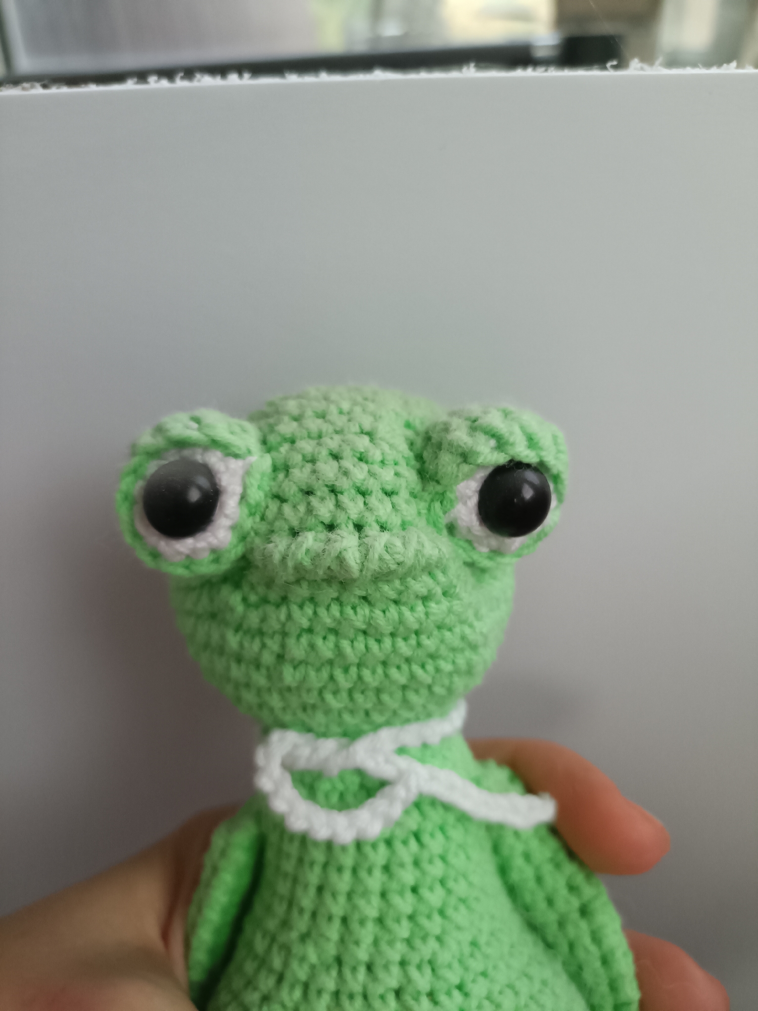A close-up, waist-up shot of a crocheted green frog toy held by a person's hand. The frog has large, black, googly eyes outlined in white, and a white crocheted bow around its neck. The background is a plain, light gray surface with a white horizontal line across the top, suggesting a wall or shelf. No specific location in Bataysk, Russia is discernible from the image.