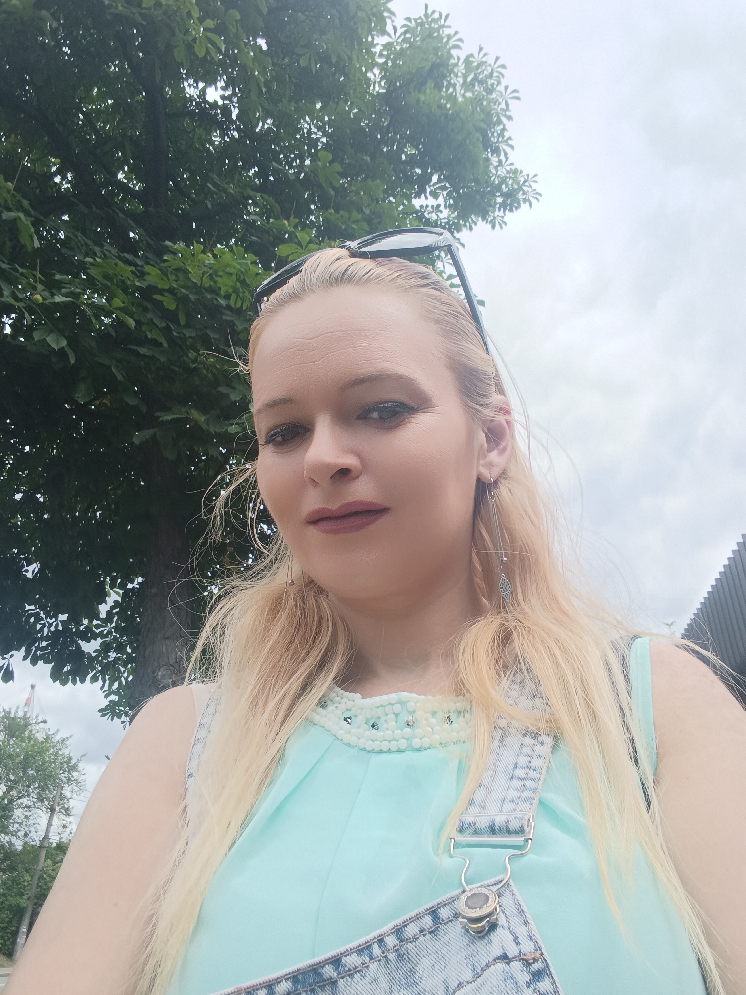 A blonde woman is pictured outdoors, likely in Chernihiv, Ukraine, as indicated by the provided information. She is wearing light blue overalls over a light blue top with pearl-like embellishments around the neckline. She has long blonde hair and is wearing long, dangly earrings. Sunglasses are perched on top of her head. The background features a large tree with green leaves and a cloudy sky. A portion of a grey, corrugated structure is visible on the right side of the image.