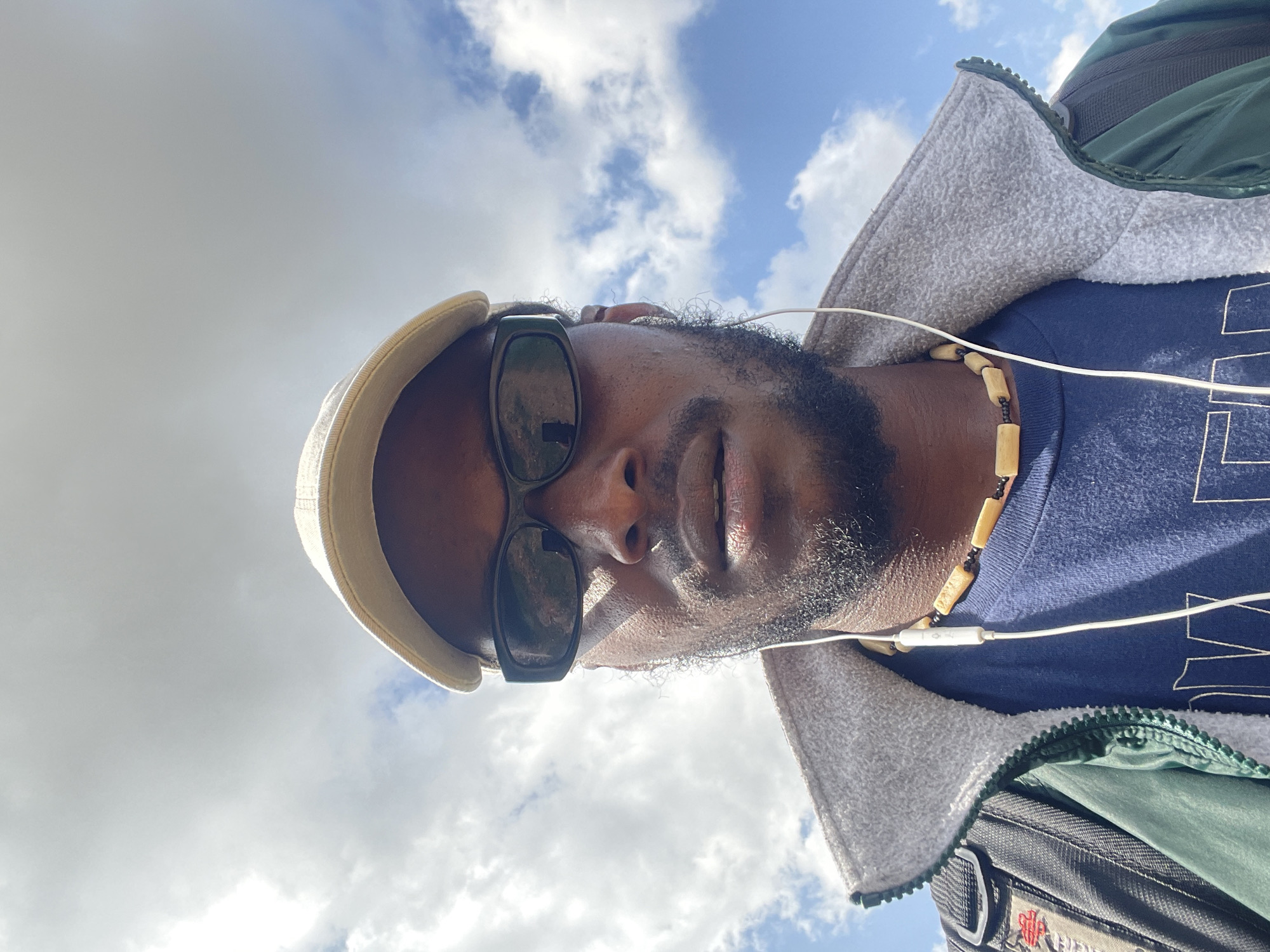 A low-angle shot captures a close-up of a Black man wearing sunglasses and a light-colored cap, with a beard and short dark hair. He is looking up towards the sky, which is filled with clouds and patches of blue. The man is wearing a dark blue t-shirt, a light gray zip-up jacket over it, and a dark green jacket partially visible. He also has a necklace made of wooden beads and a black cord, and white earbuds are in his ears. The lighting suggests it is daytime, with sunlight illuminating the scene. The background is predominantly the sky, indicating an outdoor setting. The orientation of the image is sideways, as if taken from a person lying down or looking upwards.