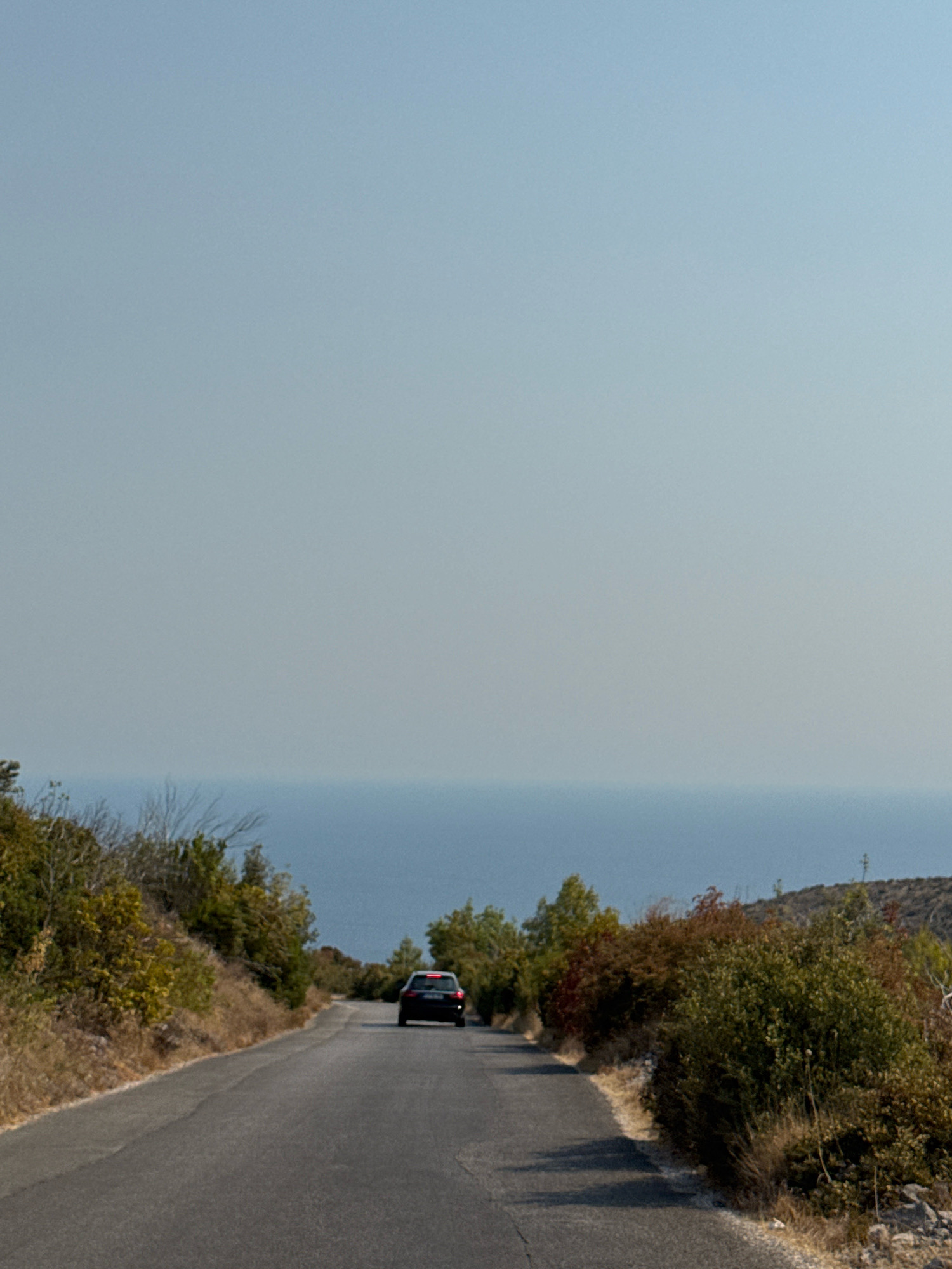 A dark-colored car drives along a paved road that curves uphill. The car is viewed from behind and is the only vehicle visible in the frame. The road is lined with dry, brown grass and dense green and reddish-brown foliage. In the background, a vast expanse of blue water, likely the sea, is visible under a pale blue sky. The scene appears to be a rural coastal area. The location is identified as Luštica, Montenegro.