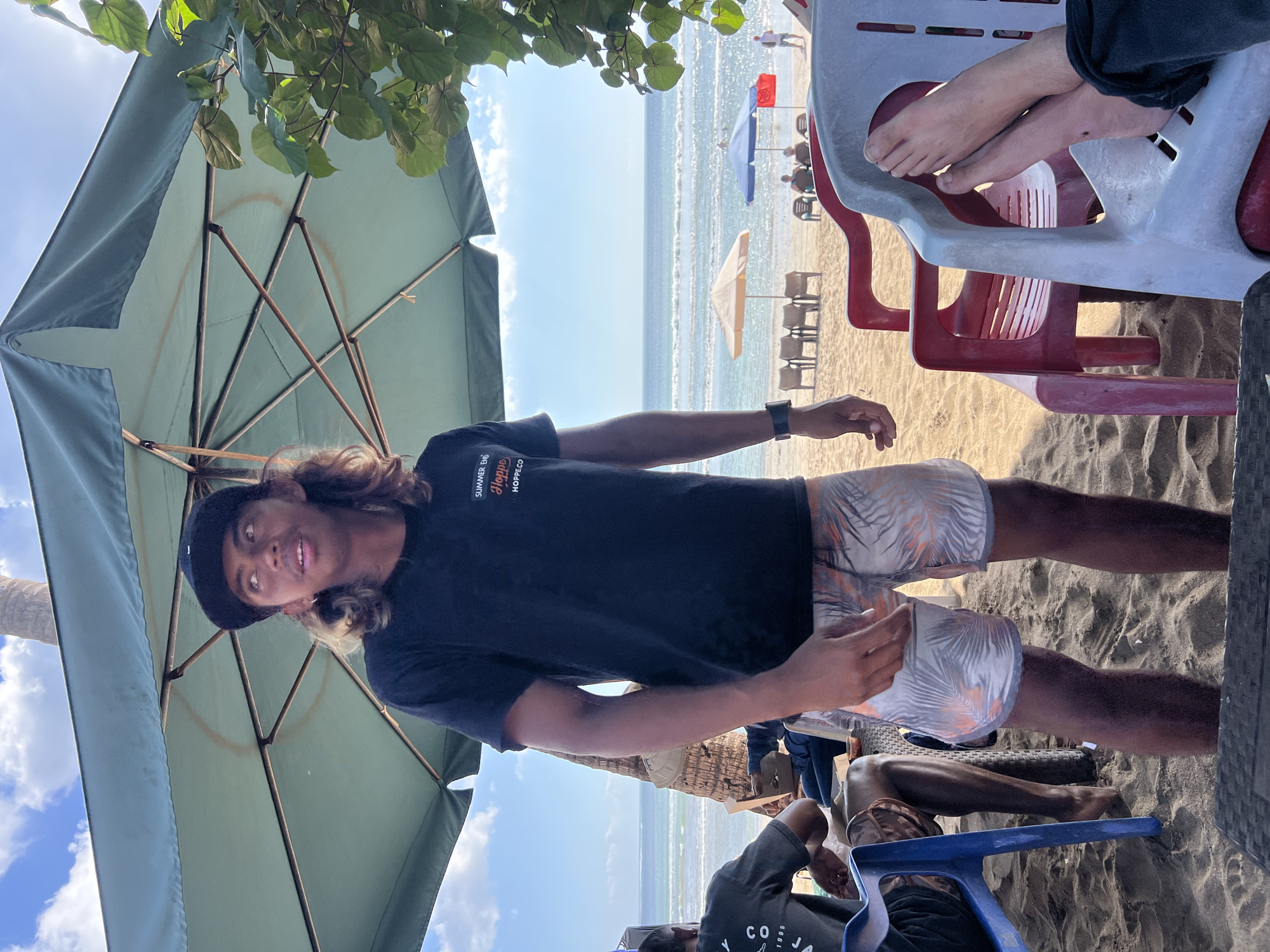 A young man with long, light brown hair and a black baseball cap is standing on a sandy beach in Kuta, Indonesia, under a large, light green umbrella. He is wearing a black t-shirt with white and orange text that reads "SUMMER END Hoppe HOPPECO.CO" and patterned swim shorts. He is looking up and to his right, with his mouth slightly open. Behind him, the ocean stretches to the horizon with gentle waves. In the background, beach chairs and umbrellas are visible on the sand, and a few people are scattered around. To the left of the young man, another person in a dark t-shirt is sitting. In the foreground, plastic chairs and a table are visible. The lighting suggests it is daytime, with a clear sky and bright sunlight.