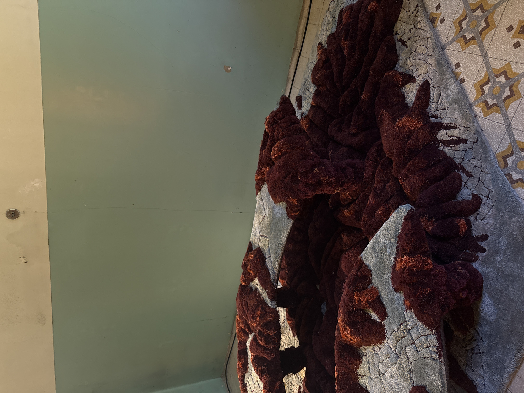 A perspective view of a room corner reveals a color palette of taupe, moss green, maroon, and gray. The wall surfaces consist of two distinct panels: a light taupe on the left and a muted moss green in the center. A circular element with a screw is noticeable on the taupe wall.
To the right, a complex, textured object dominates the view. It features maroon and gray colors and may be a rug or textile art. It has a 3D appearance with densely packed, irregular forms.
The floor at the upper right corner exhibits a hexagonal tile pattern with taupe and brown starburst designs.
The location is possibly in Milan, Italy.