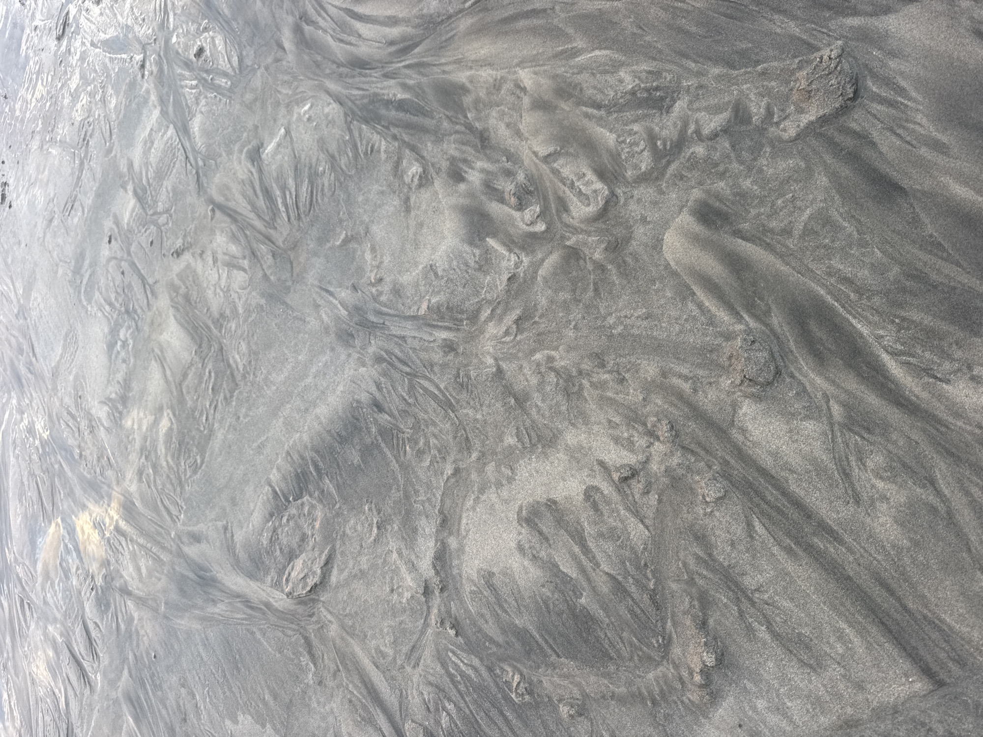 The image depicts the textured surface of wet sand on a beach, likely at low tide. The sand displays intricate patterns of ridges and channels, formed by the receding water. The coloration ranges from dark grey to light grey and white, with some areas appearing reflective due to moisture. There are no people, animals, or man-made objects visible in the image. The scene is an abstract representation of natural erosion patterns on a sandy shore. The location is Kuta Selatan, Indonesia, known for its beaches and surf spots.