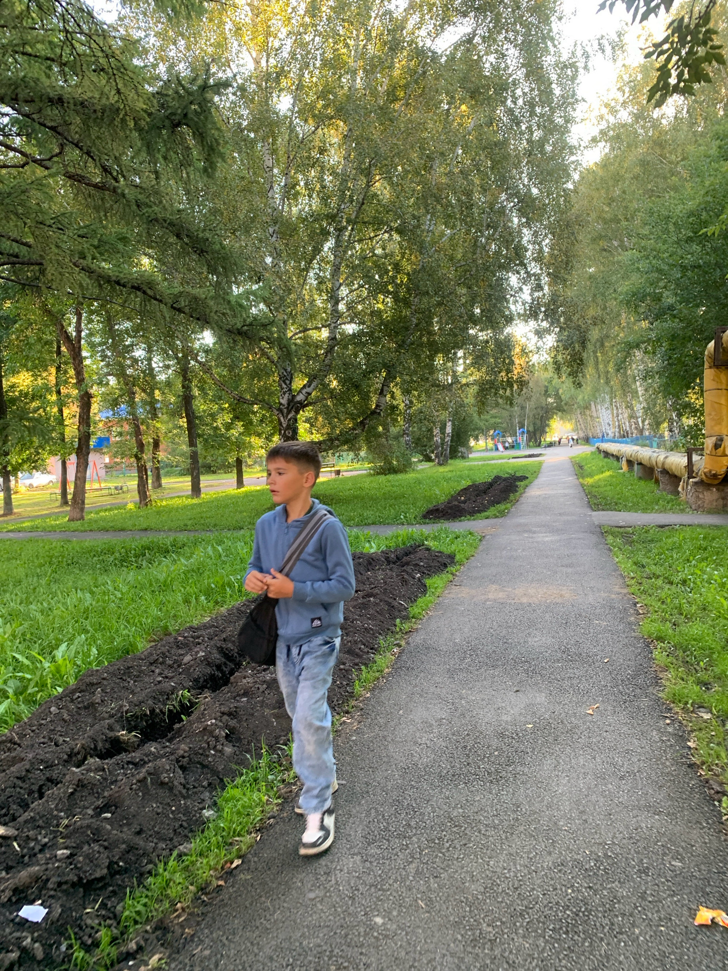 A young boy walks along a paved path lined with trees and grass. He is wearing a blue hoodie and light-colored jeans, carrying a dark bag over his shoulder. The path appears to be in a park or residential area, with a playground visible in the distance and large yellow pipes running along the right side of the path. The ground on the left side of the path is disturbed, with piles of dark soil suggesting recent landscaping work. The sunlight suggests it is either early morning or late afternoon. The location is Prokopyevsk, Russia.