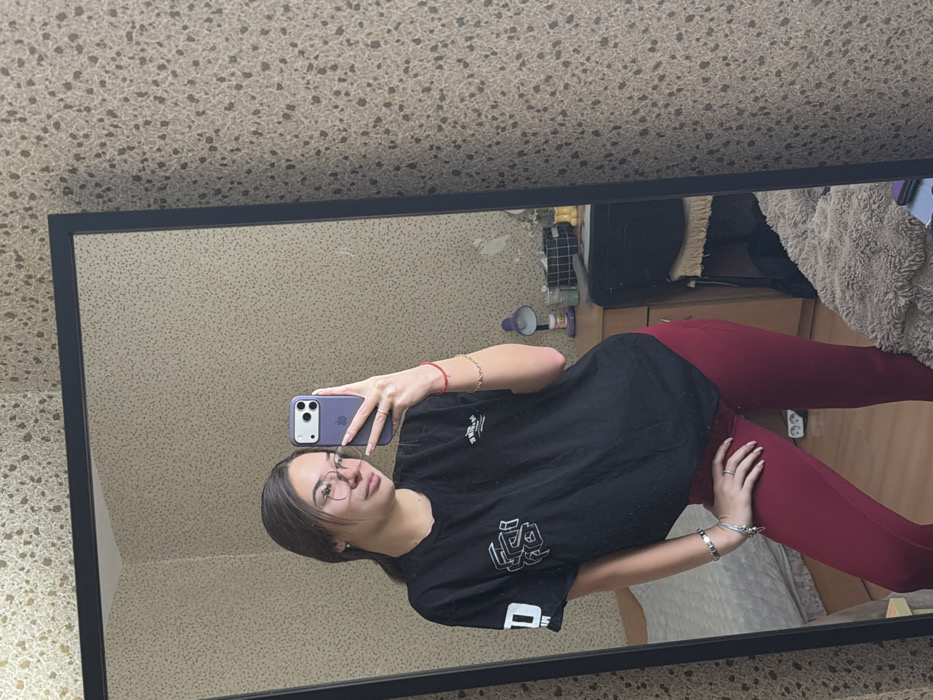 A young female subject is captured in a full-body mirror selfie taken indoors. She stands facing a large, rectangular mirror with a black frame. Her hair is dark and pulled back, and she wears clear-framed glasses. She is dressed in a black short-sleeved t-shirt featuring white graphic designs and dark red/maroon fitted leggings. Accessories include multiple silver and gold-colored bracelets on both wrists, a red string bracelet on her right wrist, and several rings on her left hand. She holds a purple-cased smartphone, likely an iPhone, in her right hand, directed towards the mirror to capture the image.
The reflection of the room visible in the mirror reveals light beige wallpaper with a dark speckled texture. To the left of the subject's reflection, a bed with a light-colored pillow and duvet is partially visible. To the right, a dark-colored bedside table or dresser holds a purple desk lamp, various small containers, and a grid-patterned basket. A fluffy, light brown blanket or rug is draped below the table. No specific visual identifiers are present within the image to determine a precise location within the city of Sofia, Bulgaria.
