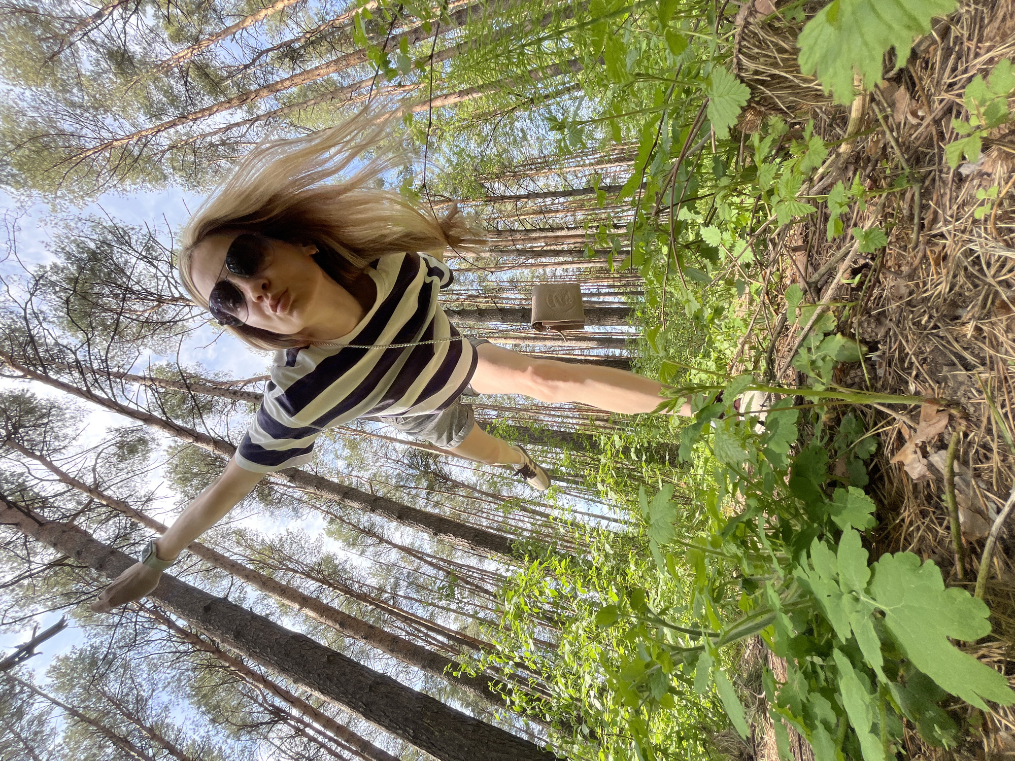 A woman wearing sunglasses, a striped top, and shorts stands in a forest in Chaikovsky, Russia. The perspective is low, looking up at her, making her appear to be standing vertically against the trees. Her blonde hair is windswept. She is wearing sneakers. The background consists of tall trees, green foliage, and a glimpse of the sky.