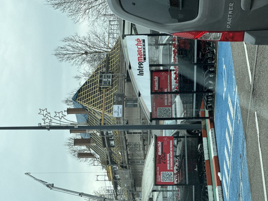 The image displays an outdoor scene, likely a commercial area, on what appears to be an overcast day. The original image is rotated 90 degrees counter-clockwise, making the horizon appear vertical. Describing it as if properly oriented:
On the left side of the frame, a building is undergoing significant construction or renovation. Its roof structure is prominently visible, showing a crisscross pattern of yellowish wooden battens over a blue tarp that covers parts of the roof. Scaffolding surrounds the building, and a white sign attached to the scaffolding reads "Wolff & Chall" and "KARNEV". In the far lower-left background, the arm of a large crane extends upwards against the pale, cloudy sky.
In front of the building, a tall black pole stands, from which a decorative element featuring three illuminated star shapes extends. Bare tree branches with no leaves are scattered across the upper-left portion of the image, suggesting winter or early spring. One tree branch appears to hold a bird's nest.
To the right of the construction site, a supermarket cart return or storage area is visible. This structure holds numerous shopping carts and features several large signs. The topmost sign is a white banner displaying "Intermarché SUPER" in red and black lettering. Below this, a red sign reads "marché d'Ober" and states "MOINS DE PAPIER, ET TOUJOURS AUTANT DE PROMOS." (less paper, and always as many promotions) along with a QR code. Another similar red sign is partially visible below it, also containing text and a QR code.
On the far right of the image, the rear passenger side of a grey van or car is visible. The word "PARTNER" is printed in white letters on its side, and a red taillight is clearly seen. The ground in the immediate foreground, to the right, is part of a parking area. A section of the ground is painted bright blue with white markings, possibly indicating a designated parking spot. An orange and white striped curb or barrier separates this blue area from other ground surfaces.