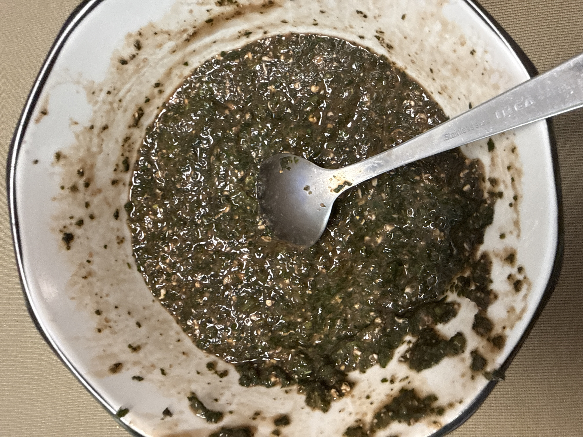 A close-up shot shows a white bowl with a black rim containing a dark green, watery substance. A silver spoon rests in the bowl, the handle displaying the inscription "Stainless Steel IKEA." The bowl's interior shows splatters and remnants of the substance clinging to the sides. The bowl is placed on a beige surface. The location is not discernible from the visual information. There is no information about people, actions, interactions, or the environment.