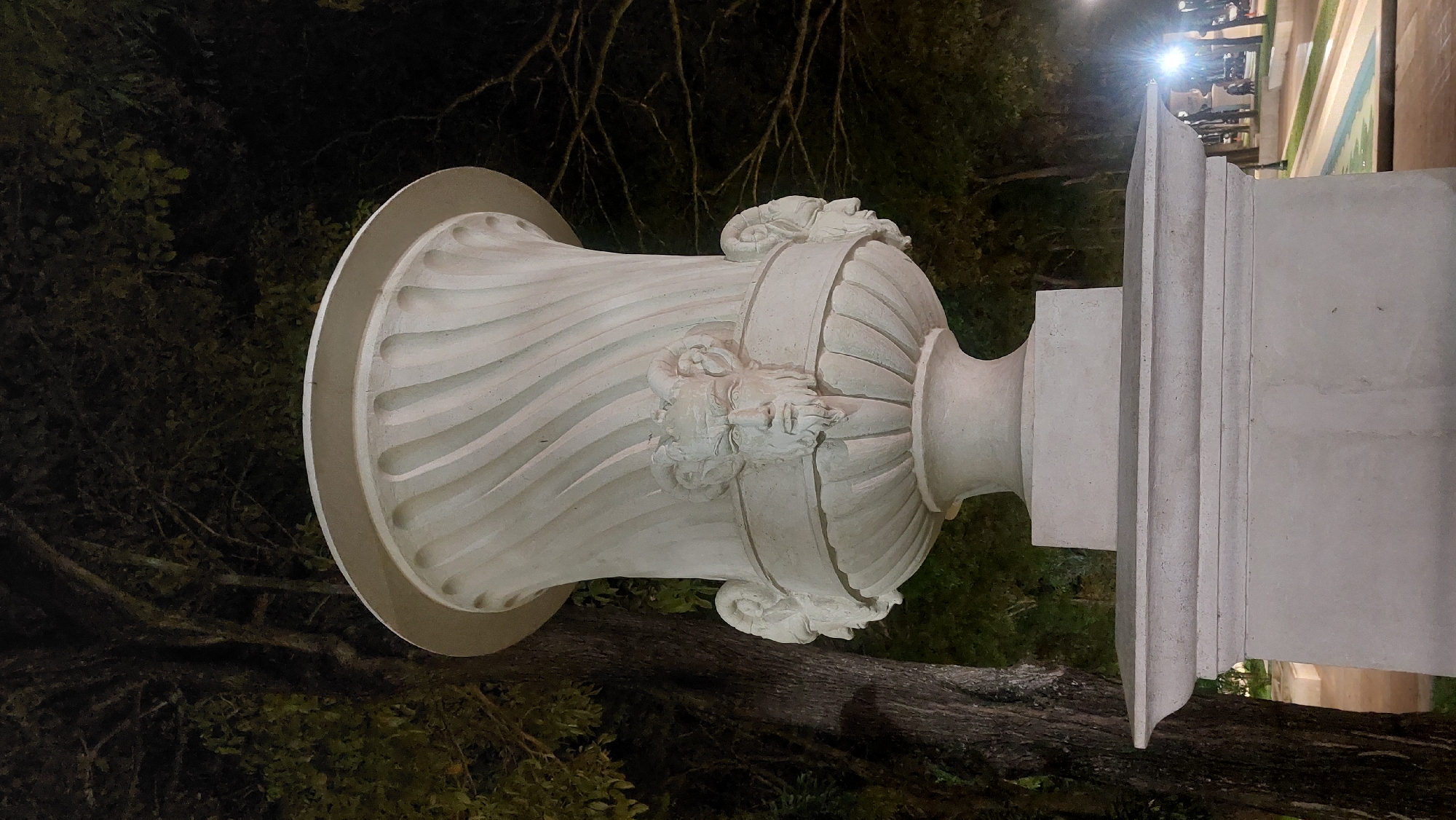 The image shows a white, sculpted urn-shaped structure outdoors. The urn is decorated with sculpted heads, situated upon a square pedestal with a series of tiered layers at the top. The background features foliage, including what appears to be a tree trunk and branches, with city lights dimly visible. Based on the prompt, the location is presumed to be Caracas, Venezuela. The image seems to be taken at night or in low-light conditions. There are no discernible people in the immediate scene.