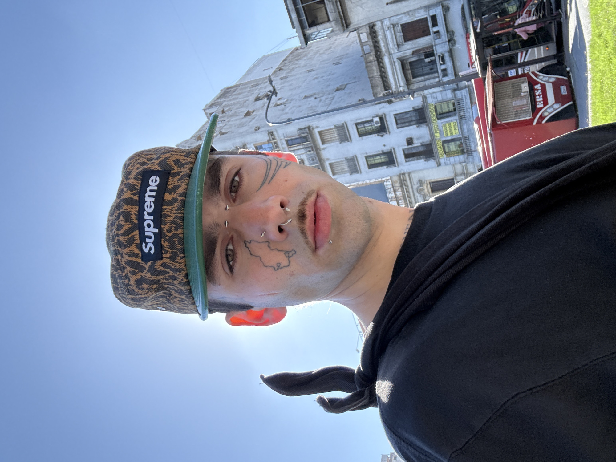 A close-up, slightly distorted, tilted shot of a young man wearing a leopard print Supreme cap with a green brim, piercings on his eyebrow, nose, and lip, and facial tattoos (one resembling a map). He is wearing a black hoodie. The background reveals an urban environment. Architectural details indicate buildings in Buenos Aires, Argentina. The red "BERSA" kiosk and building style specifically suggest the city center area. The sky is clear and blue, indicating daylight. The image seems to have been taken from a low angle, looking up at the subject.