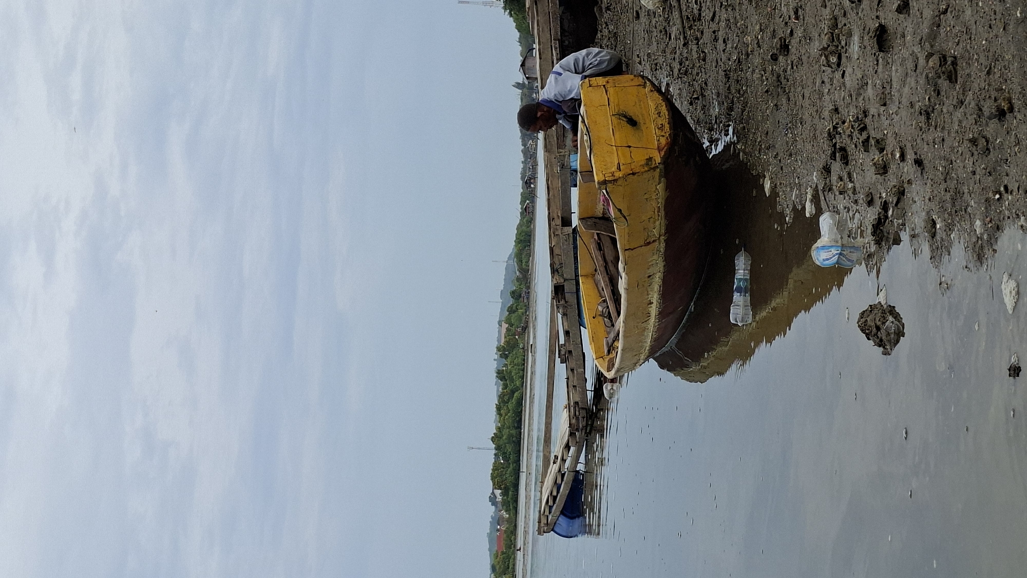 A man wearing a light blue collared shirt and dark pants crouches by the side of a weathered, yellow and red wooden boat pulled onto a muddy shore. The boat is positioned at an angle, with its bow facing left and its stern to the right. In the background, a wooden pier extends into the water, supported by blue barrels. The water is calm and reflects the overcast sky. Across the water, a distant shoreline is visible, with scattered buildings and lush green trees. Two plastic water bottles are present on the muddy bank near the water's edge. The scene appears to be a coastal area in Lhokseumawe, Indonesia.