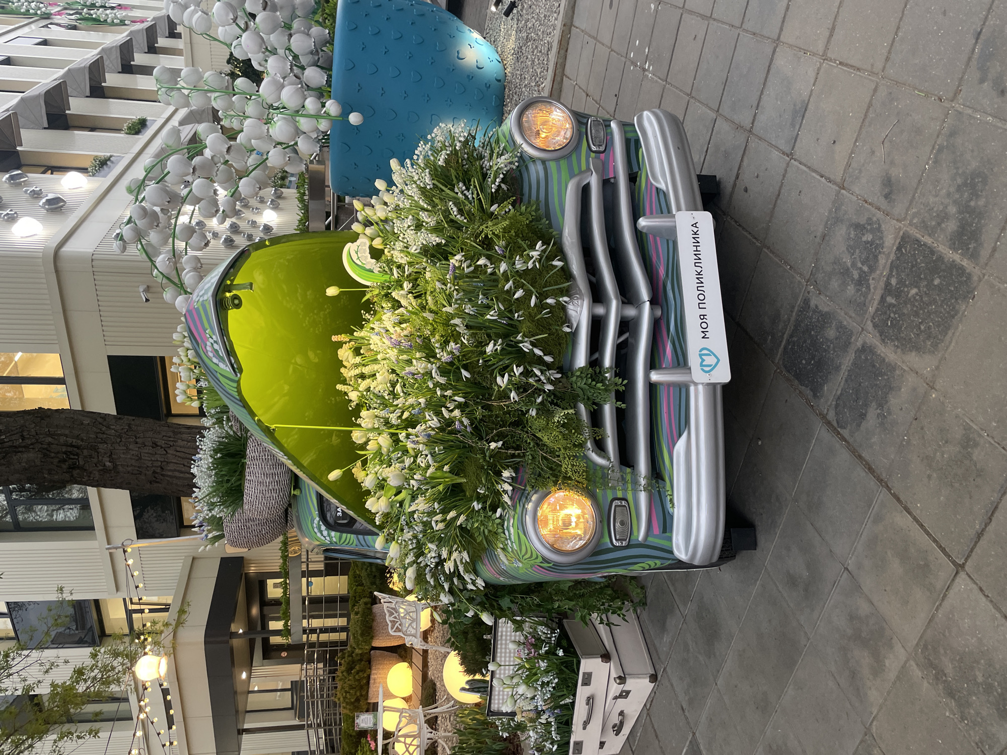 A vintage car decorated with an abundance of white flowers is parked on a brick sidewalk. The car's hood and front grill are covered in the floral display. A sign with Cyrillic text is affixed to the front of the car. The car's headlights are illuminated.
The background features modern buildings with visible windows and awnings. Streetlights and other decorative lighting are also visible in the surrounding area.
Based on the Cyrillic text and architectural style, the image is likely taken in Moscow, Russia. The scene appears to be a decorated street or public space, possibly for a special event or promotion.