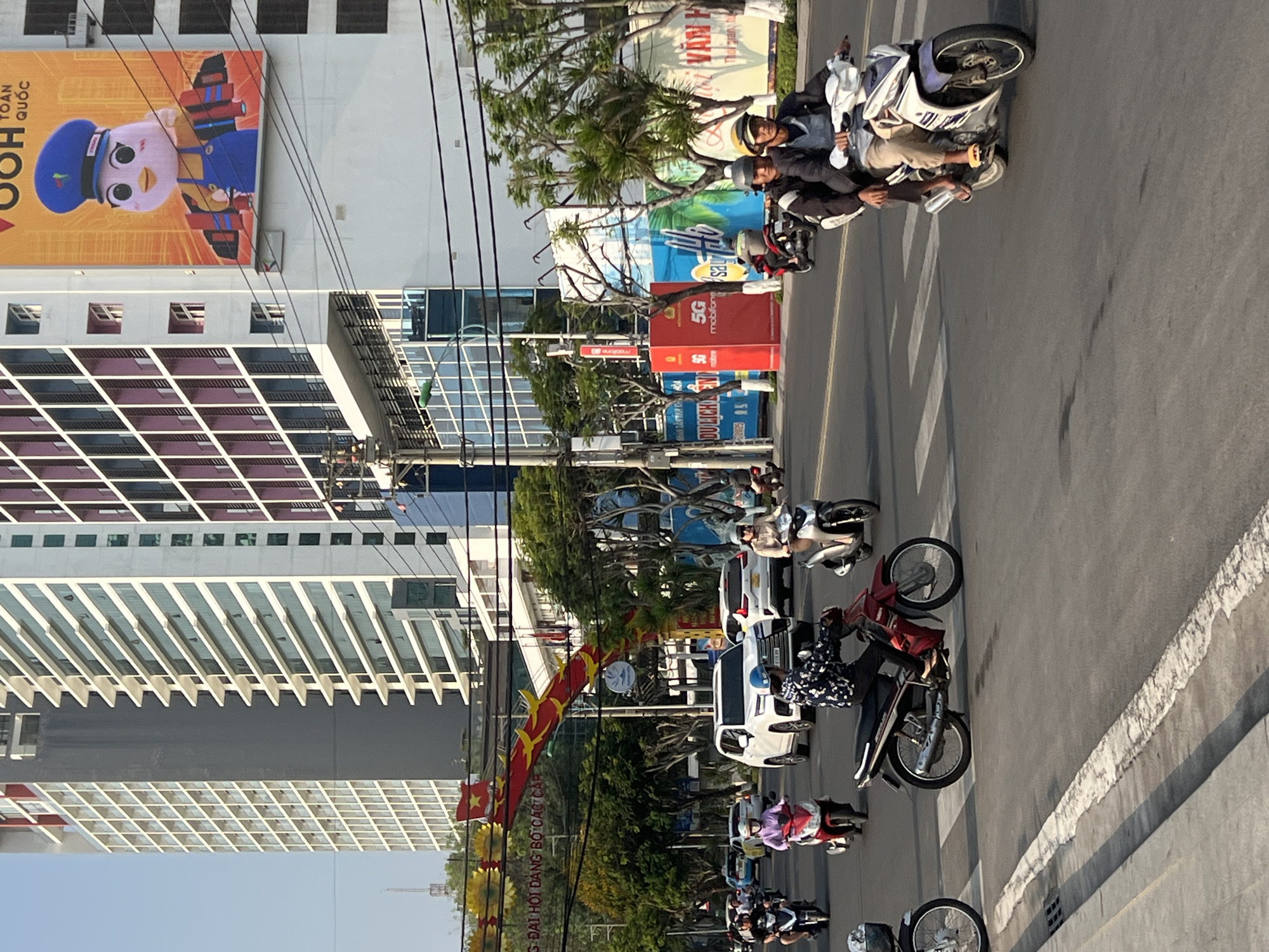 The image shows a street scene in Nha Trang, Vietnam, with a tall building on the left side of the frame featuring a large advertisement of a cartoon character dressed as a postal worker in a blue uniform. Various buildings are behind trees and street lights, one with an advertisement of the number 5G. A red banner with orange flags, resembling a dragon, stretches across the road. Several motorcycles, some carrying multiple people, are traveling along the road, and a white car is visible in the middle of the road. The sky is light blue, suggesting a sunny day.