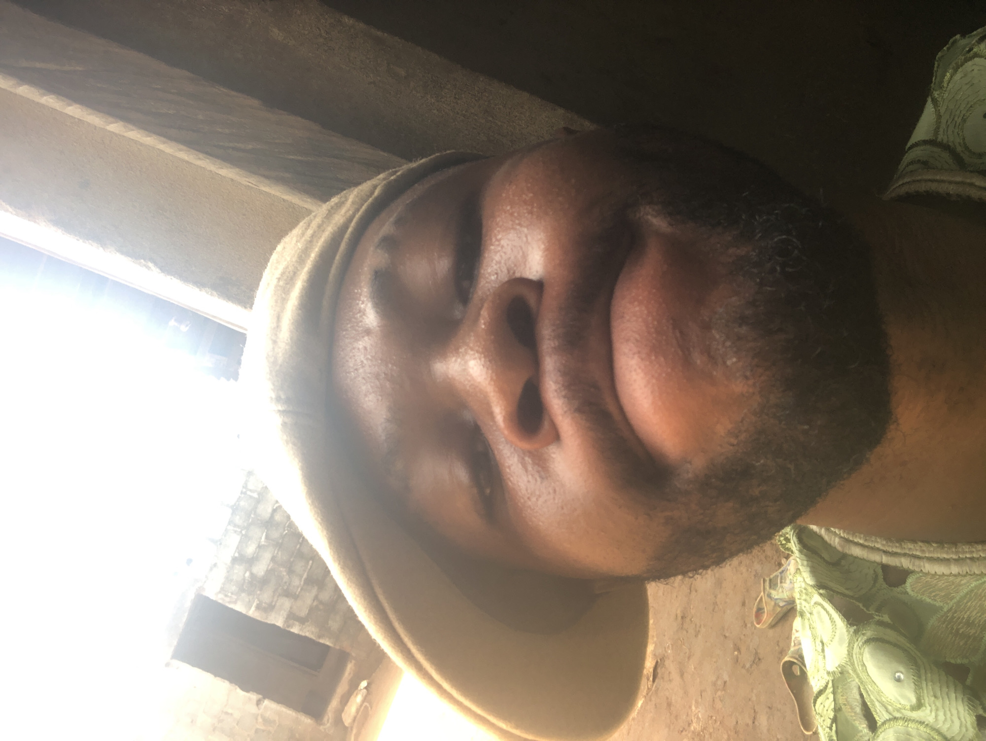 A close-up, rotated portrait image features a dark-skinned man with a beard. The image is oriented as if viewed from an upward angle, with the man's head tilted. He wears a light brown or beige cap. His facial expression shows his mouth pursed or shaped into a distinct pout, and his visible eye appears to be squinting or partially closed.
A strong, overexposed light source, likely sunlight, streams in from the left side of the frame, creating harsh highlights on his face and blowing out details in the background. Within this bright area, some rough, grey brickwork can be discerned, suggesting an opening like a window or doorway. The rest of the immediate background above him is dark and shows a textured, possibly concrete or wooden, surface.
On the right side of the image, part of a green garment with an intricate pattern is visible. The lighting conditions suggest it is daytime. There is no discernible text in the image. The location appears to be indoors or under a simple shelter, possibly an unfinished structure given the brickwork. The overall impression is a somewhat quirky or playful selfie.