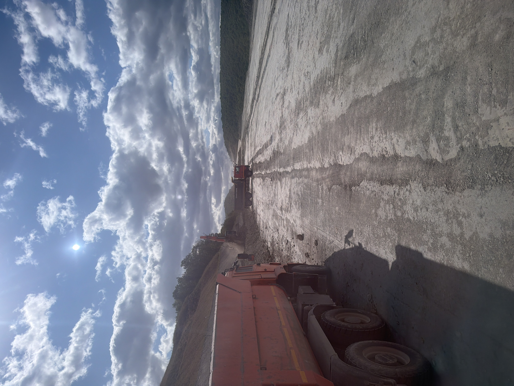 An orange dump truck is parked on a wide, dirt road. In the background, an excavator is digging into a hillside. Another dump truck with "SINOTRUK" written on it is further down the road. The sky is blue with white and gray clouds, and the sun is shining brightly. The scene appears to be a construction site in the city of Kolotov Buerak, Russia.